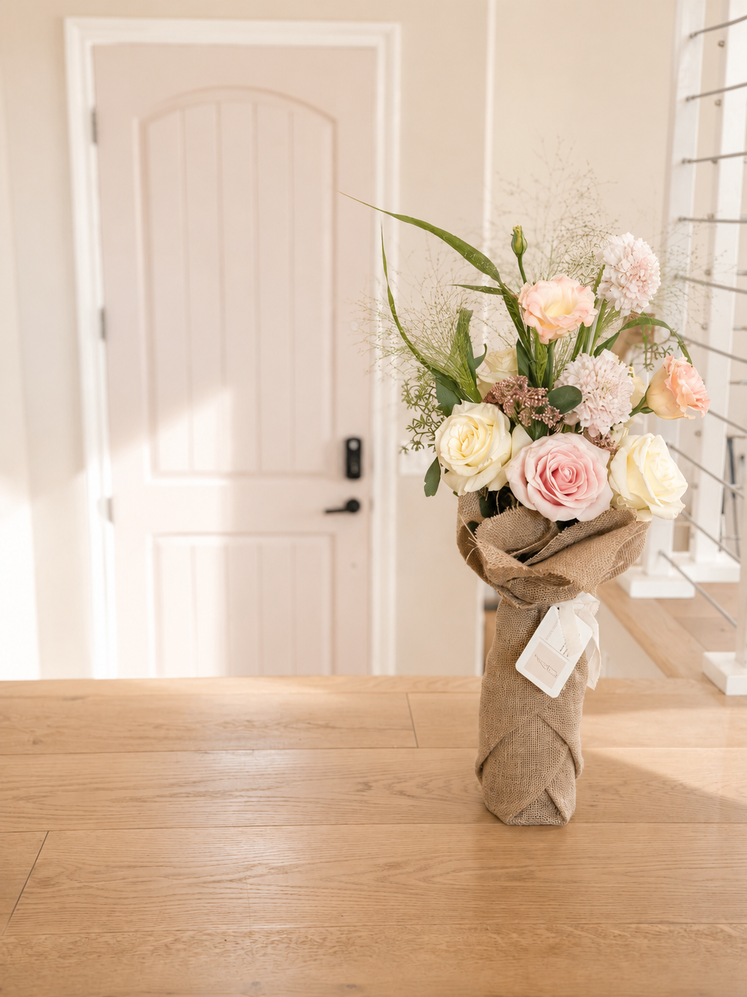 A bouquet of pink and white roses and hydrangeas in a burlap-wrapped vase on a wooden table in a room with a door and a window.