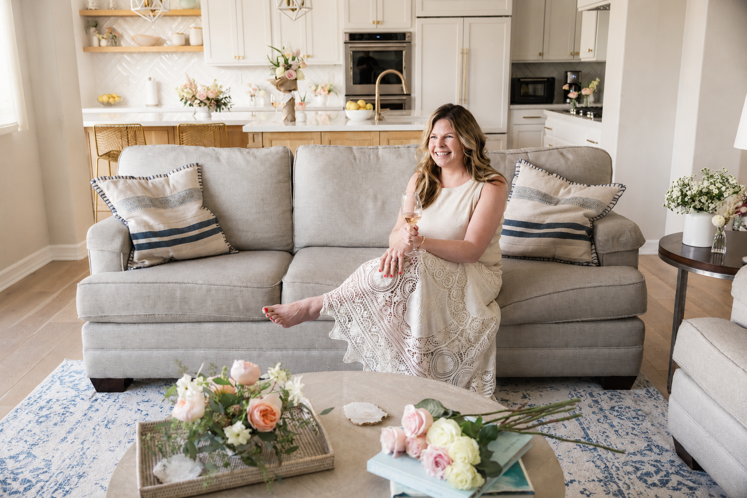 A woman sitting on a beige sofa with floral pillows, holding a glass of white wine, in a bright living room with a kitchen in the background and a bouquet of flowers on the coffee table.