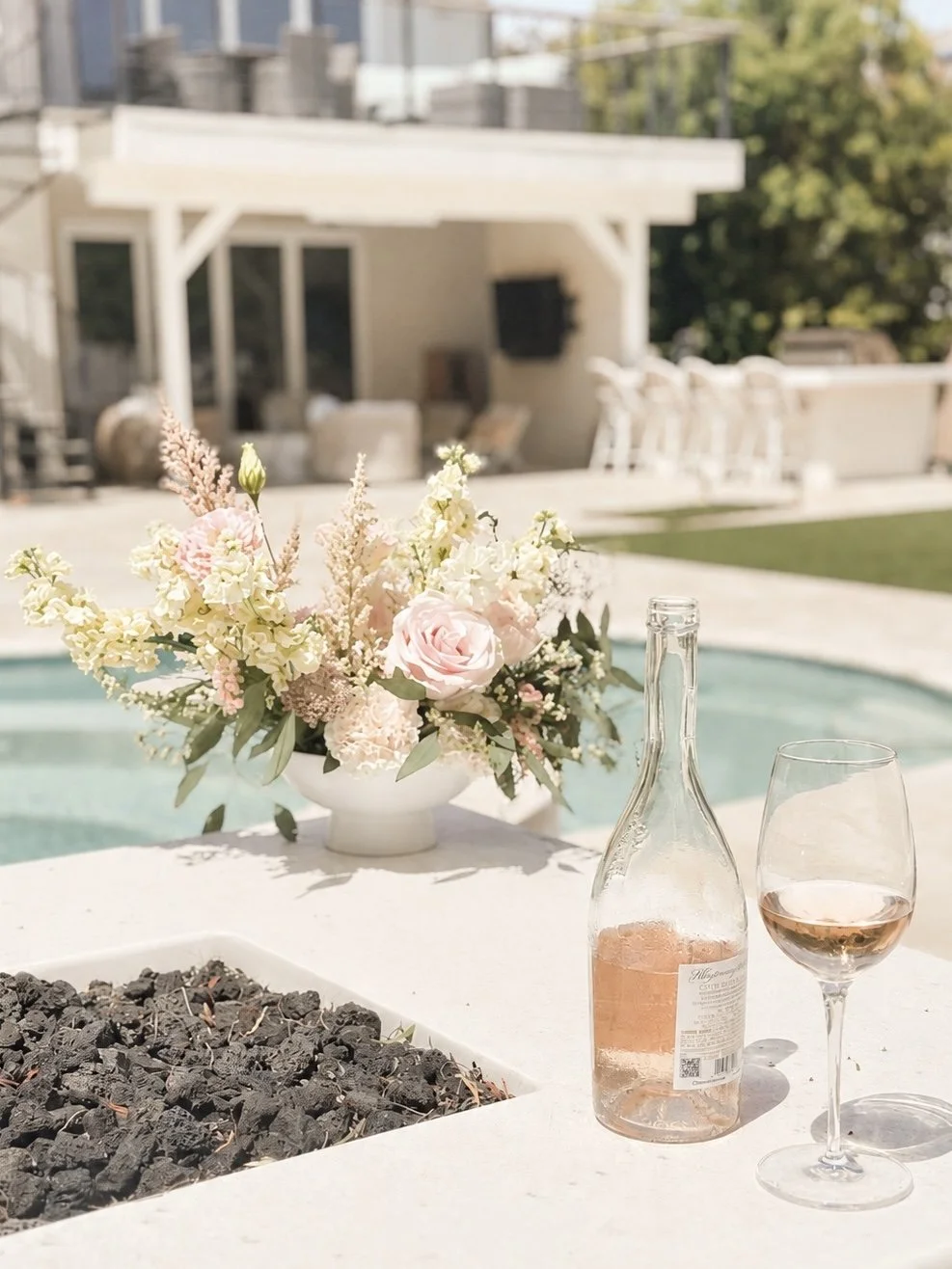 A table with a pink wine bottle, a glass of rosé, a white flower arrangement, and a fire pit, set outdoors near a swimming pool.