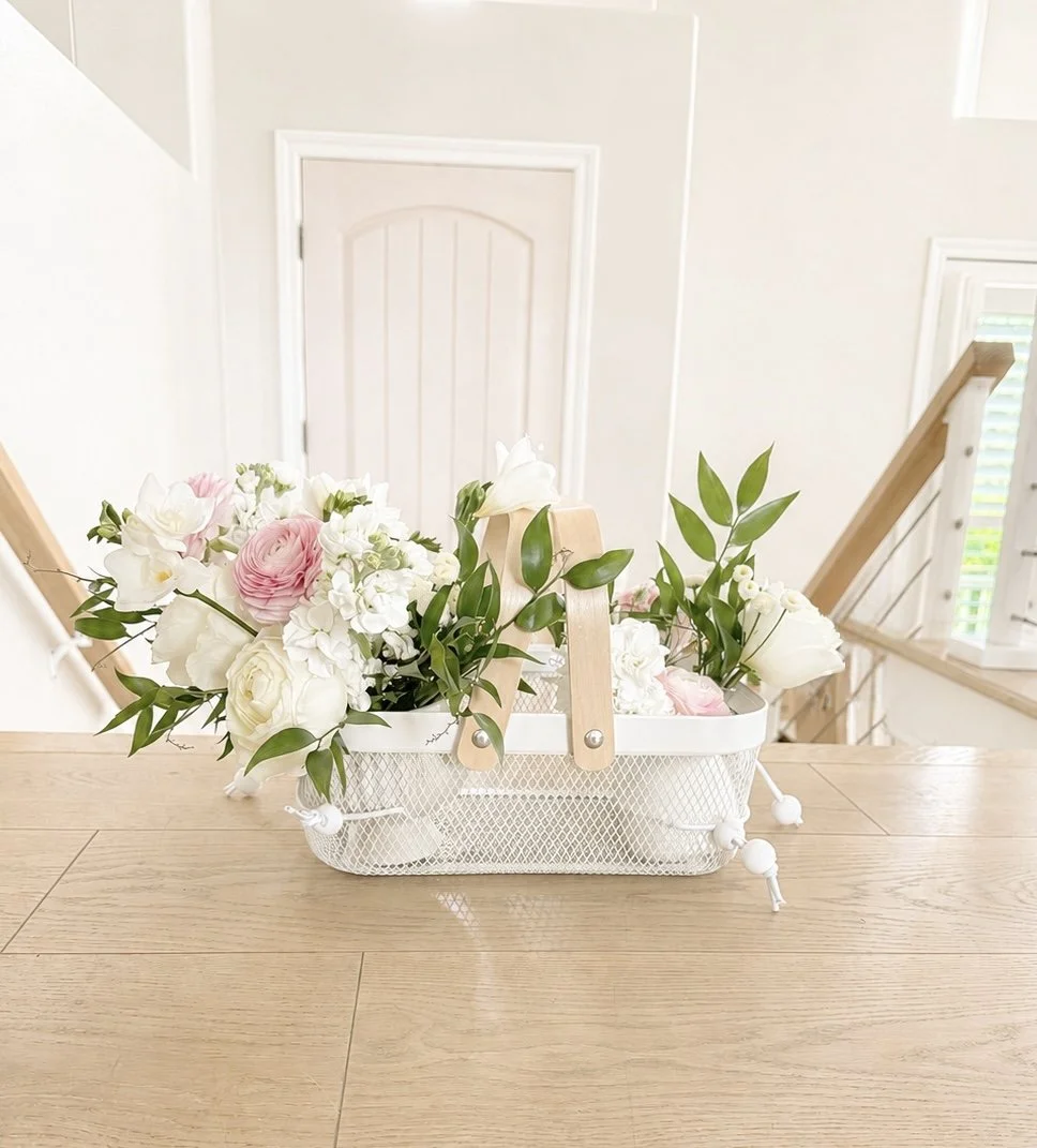 A white basket with a wood handle filled with pink and white flowers and green leaves, placed on a wooden table in front of a staircase and a closed door in a bright home.