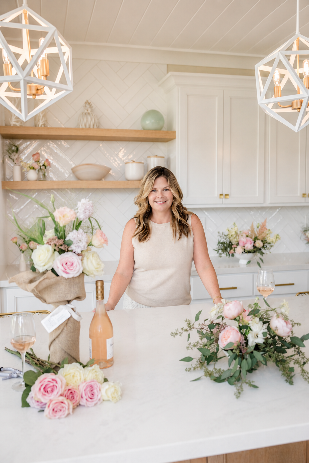 A woman smiling at a kitchen island decorated with pink and white flowers, a bottle of rosé wine, and two glasses of rosé wine.