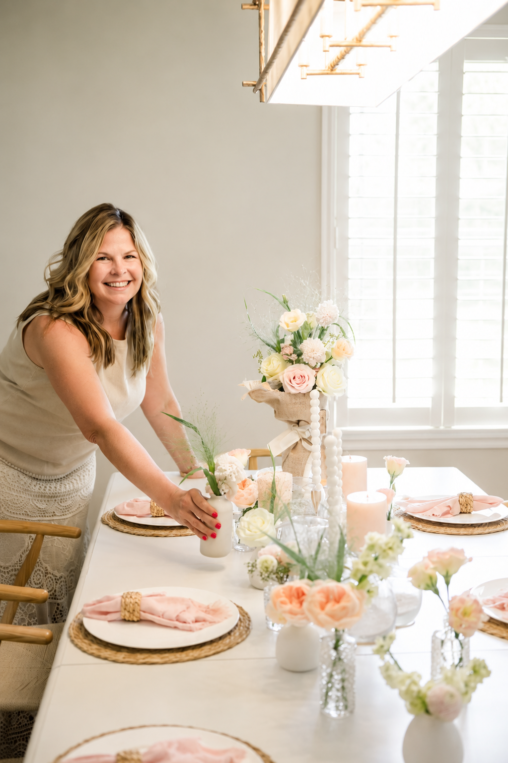 Woman smiling and arranging pink and white flowers on a decorated dining table with candles and place settings near a window with white shutters.