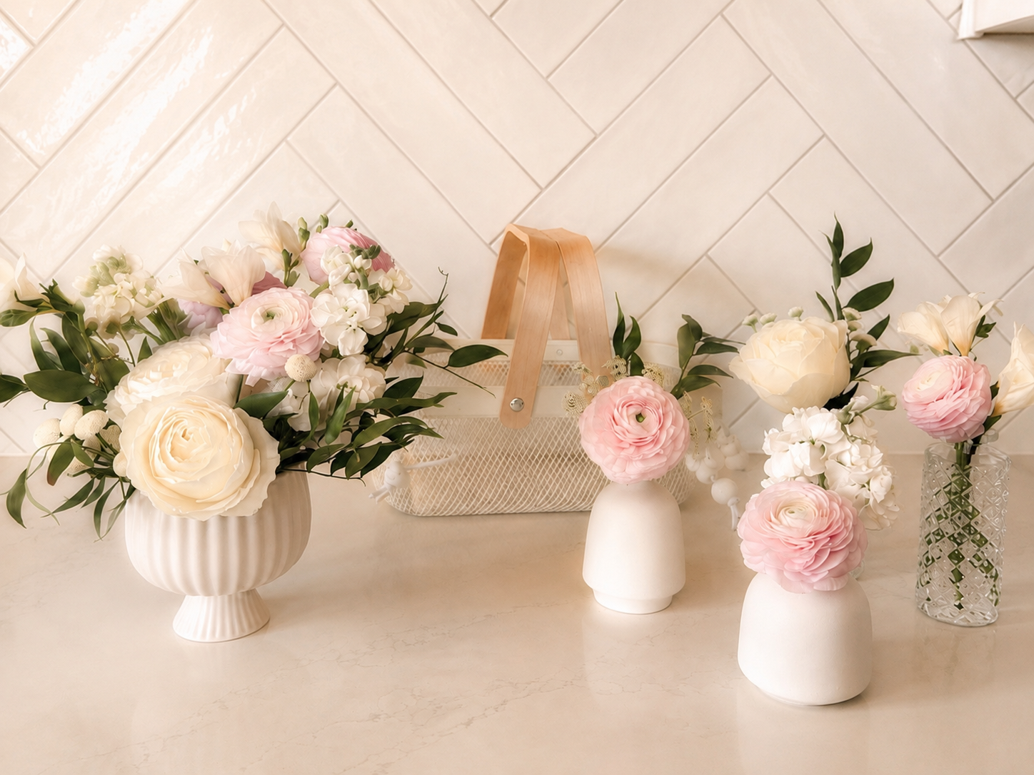 Four vases with white and pink flowers arranged on a cream-colored surface, with a beige textured tiled wall in the background.