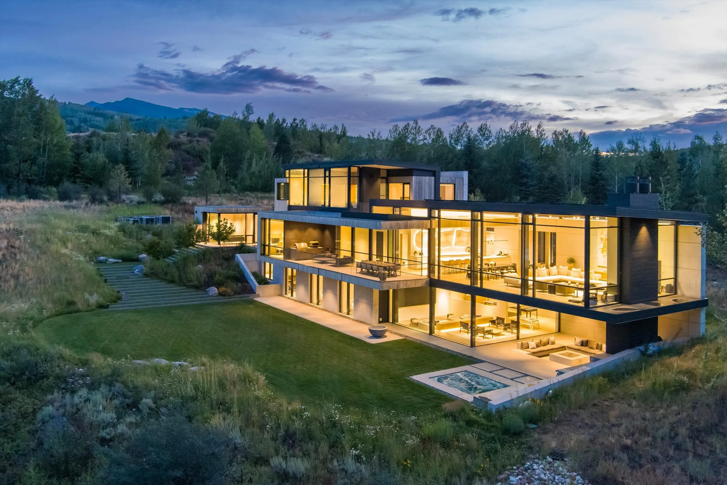 Modern multi-story house with large glass windows at dusk, illuminated from inside, surrounded by greenery and hills in the background.