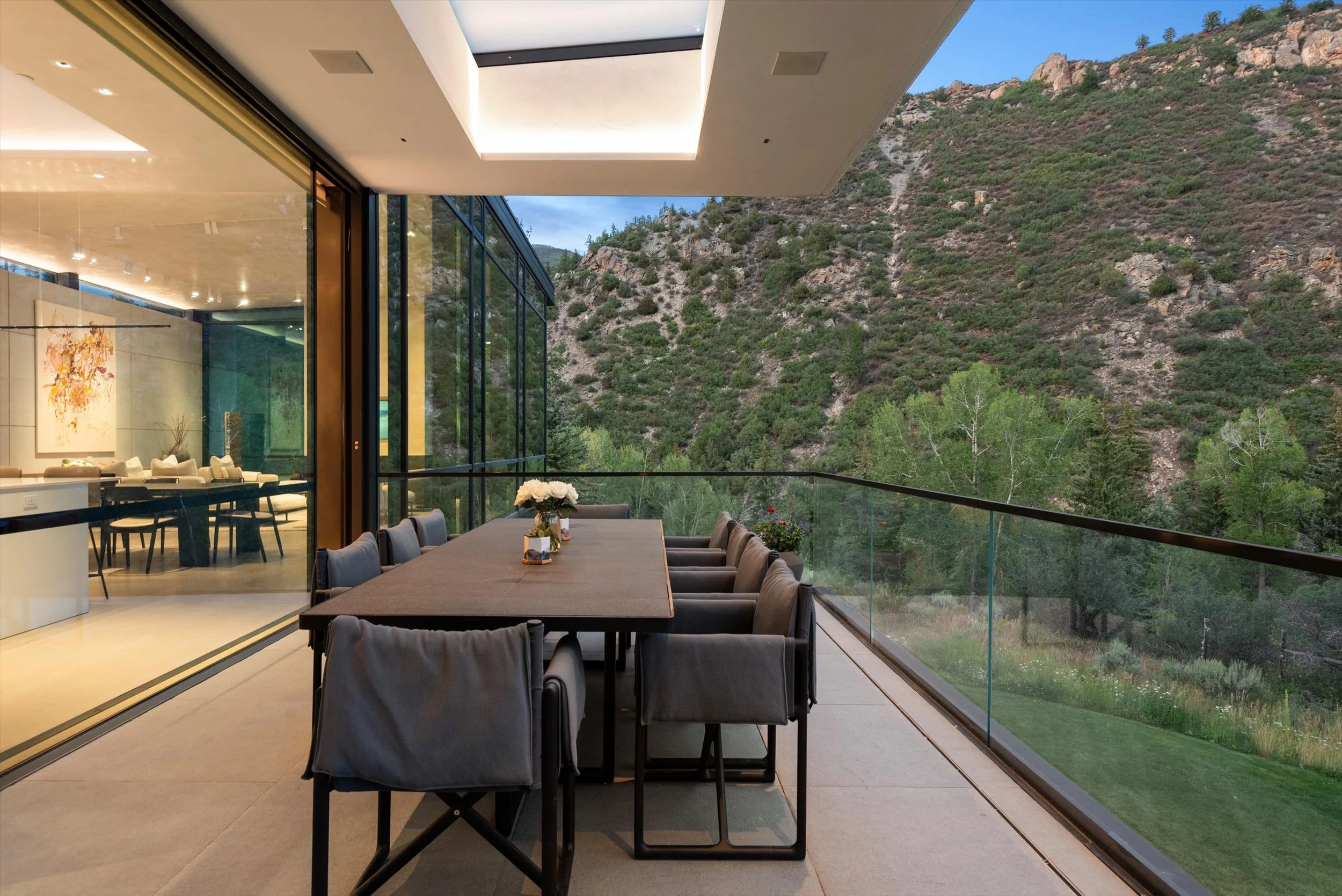 A spacious outdoor patio with a large dining table and chairs, overlooking a forested hillside with rocky terrain.