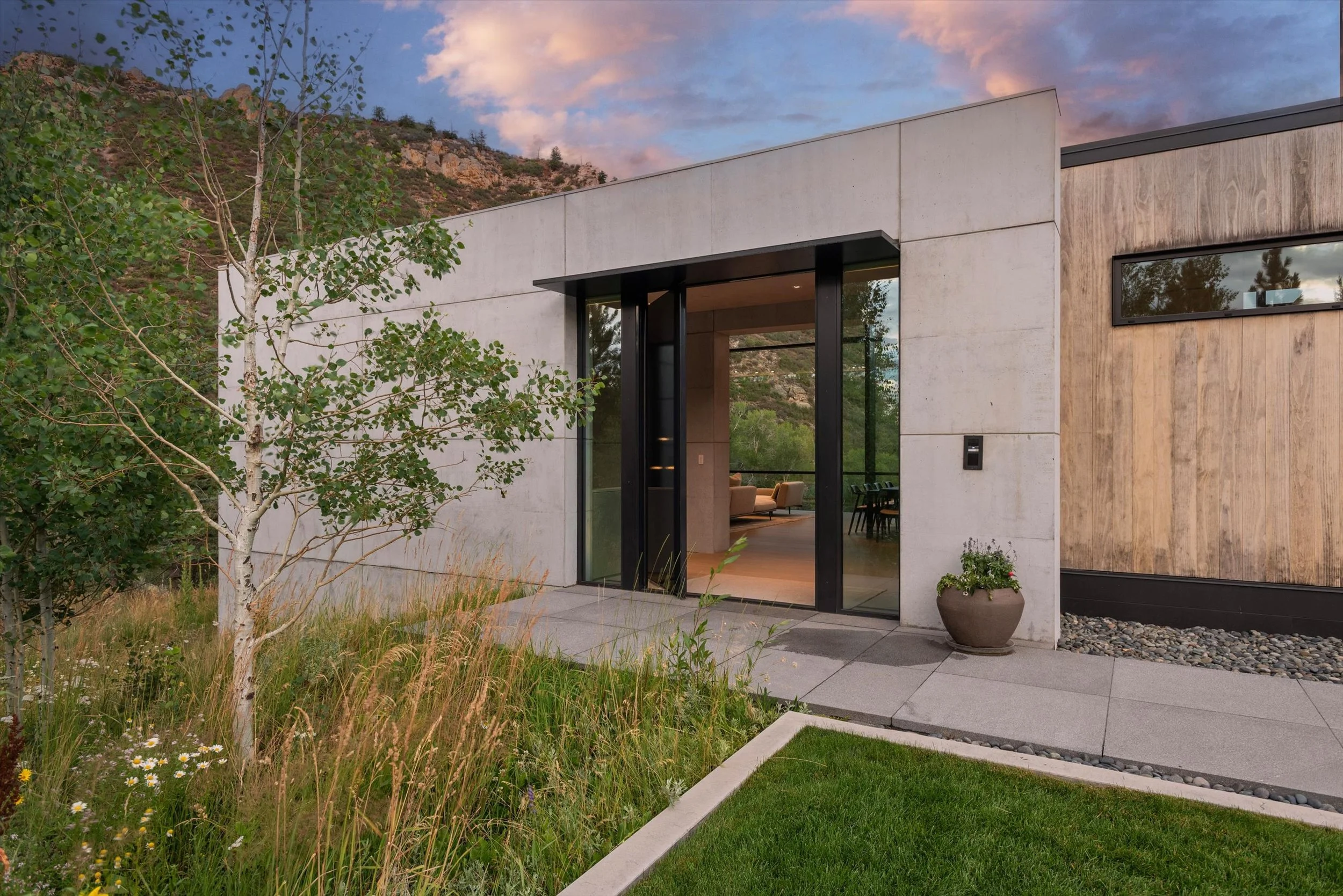 Modern house with large glass doors, surrounded by greenery and mountain landscape, during sunset.