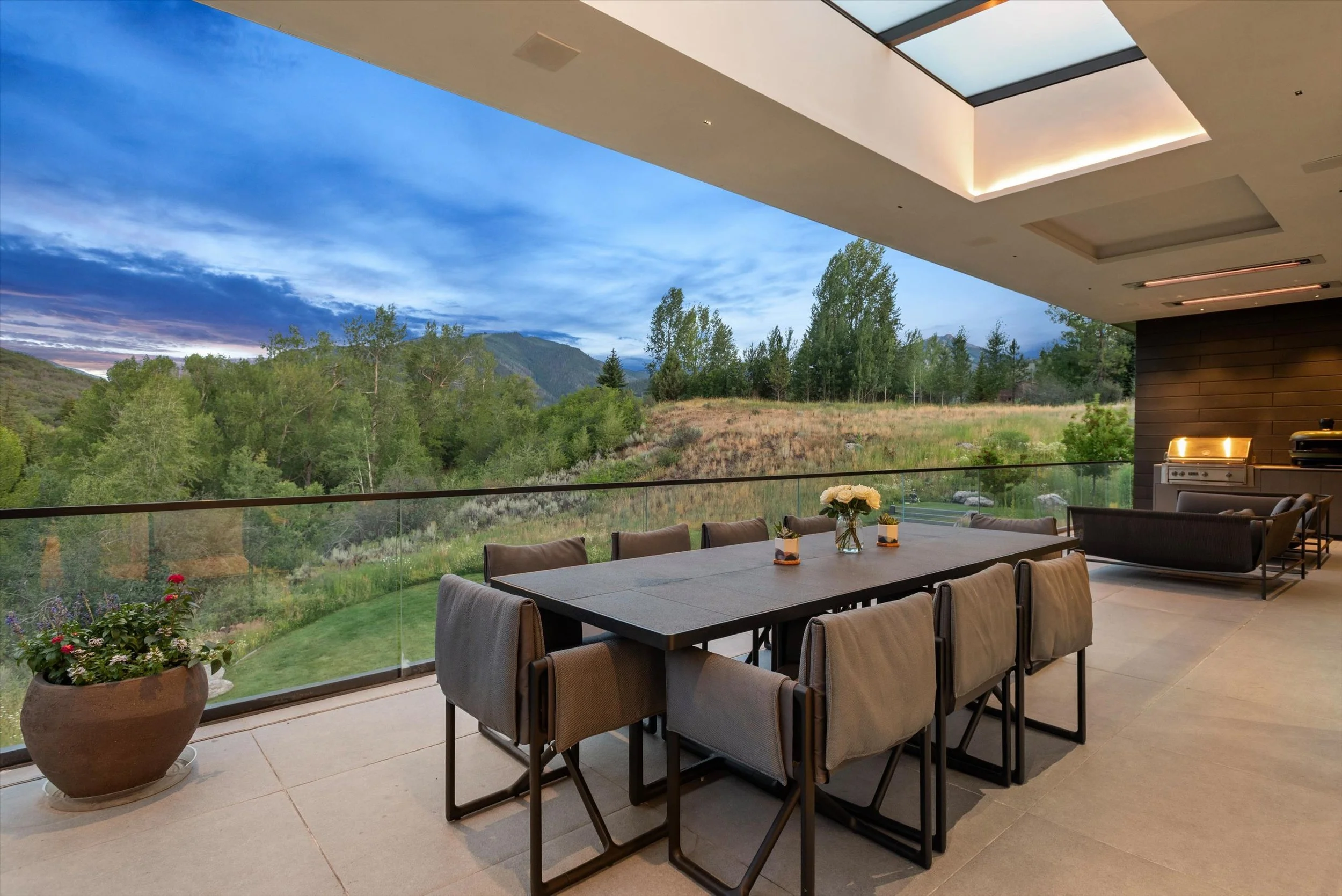 Modern outdoor patio with a dining table, chairs, and a view of green trees and mountains under a partly cloudy sky, with a built-in outdoor kitchen area.
