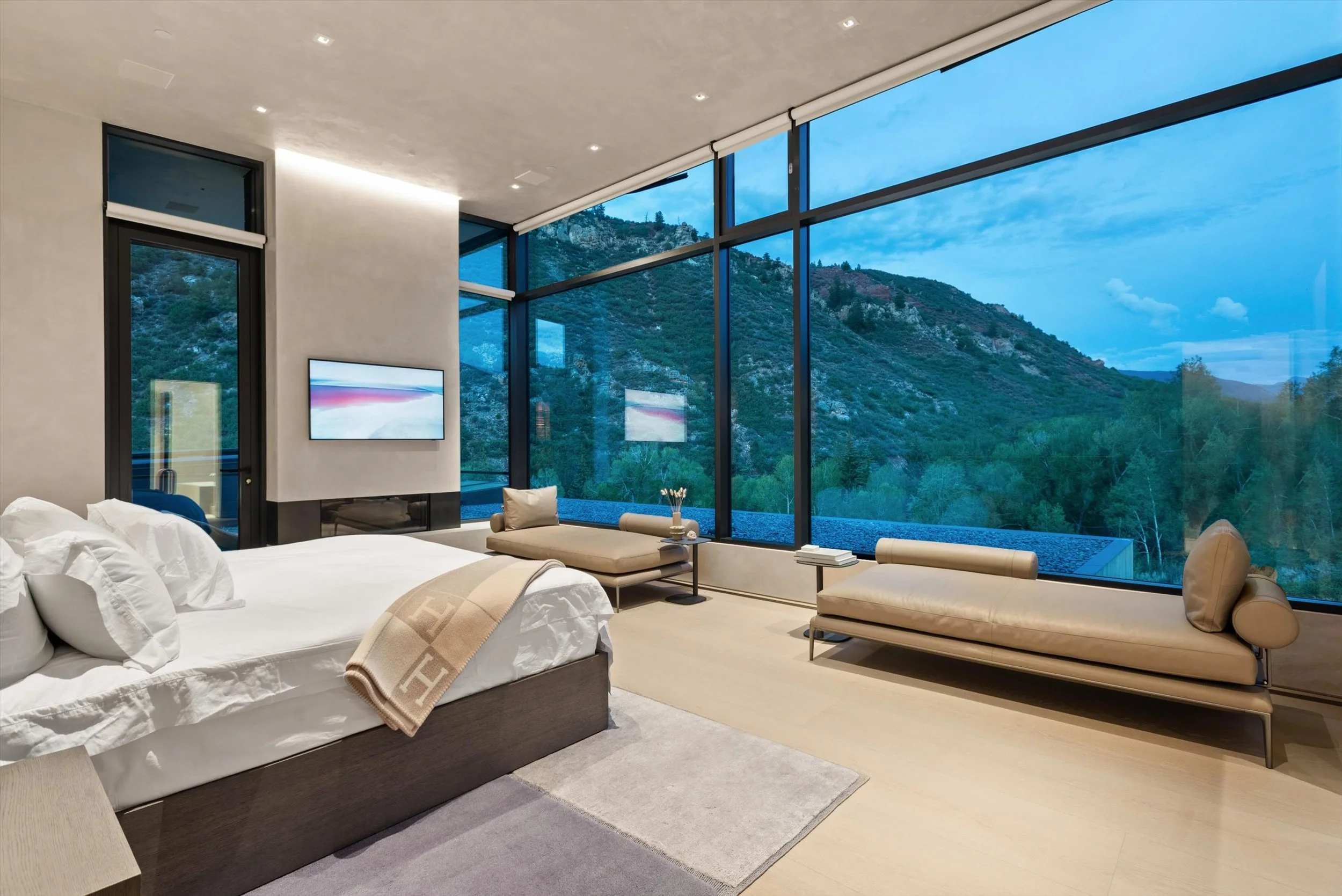 Modern bedroom with large floor-to-ceiling windows overlooking a mountain landscape, featuring a white bed, beige lounge chairs, and a television mounted on the wall.