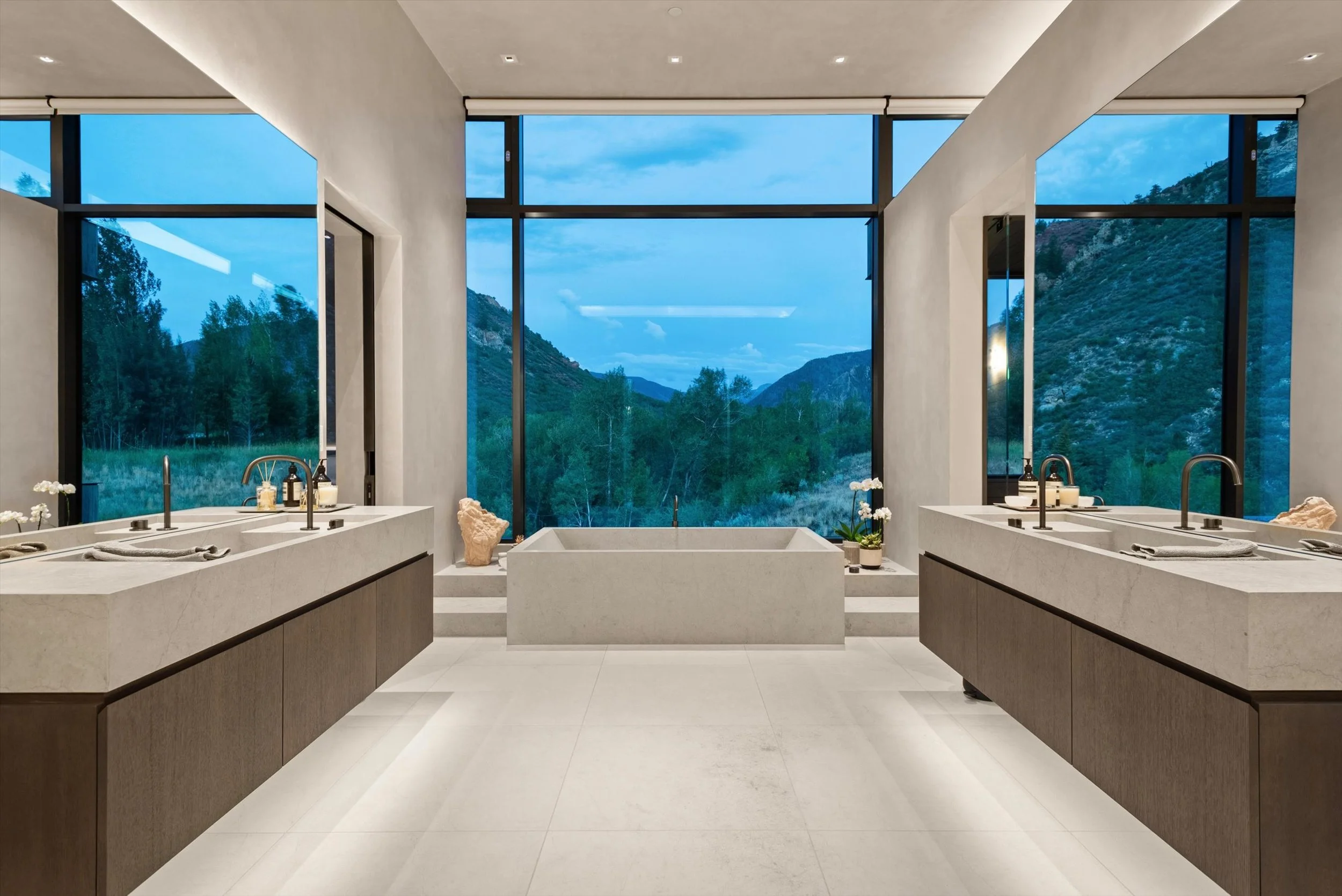 Modern bathroom with large floor-to-ceiling windows showcasing mountainous landscape, featuring two symmetrical sinks with soap dispensers and towels, decorative rocks, and potted plants.