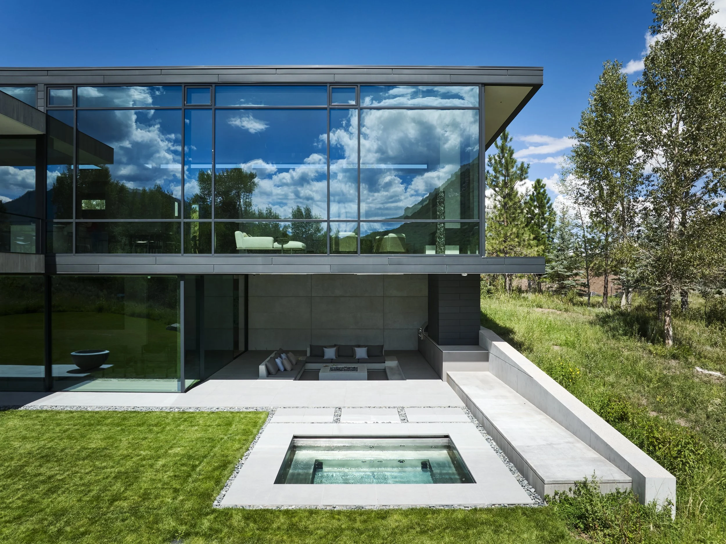 Modern house with large glass windows reflecting the sky and trees, patio with outdoor seating, small pool or hot tub, surrounded by grass and trees under a partly cloudy sky.