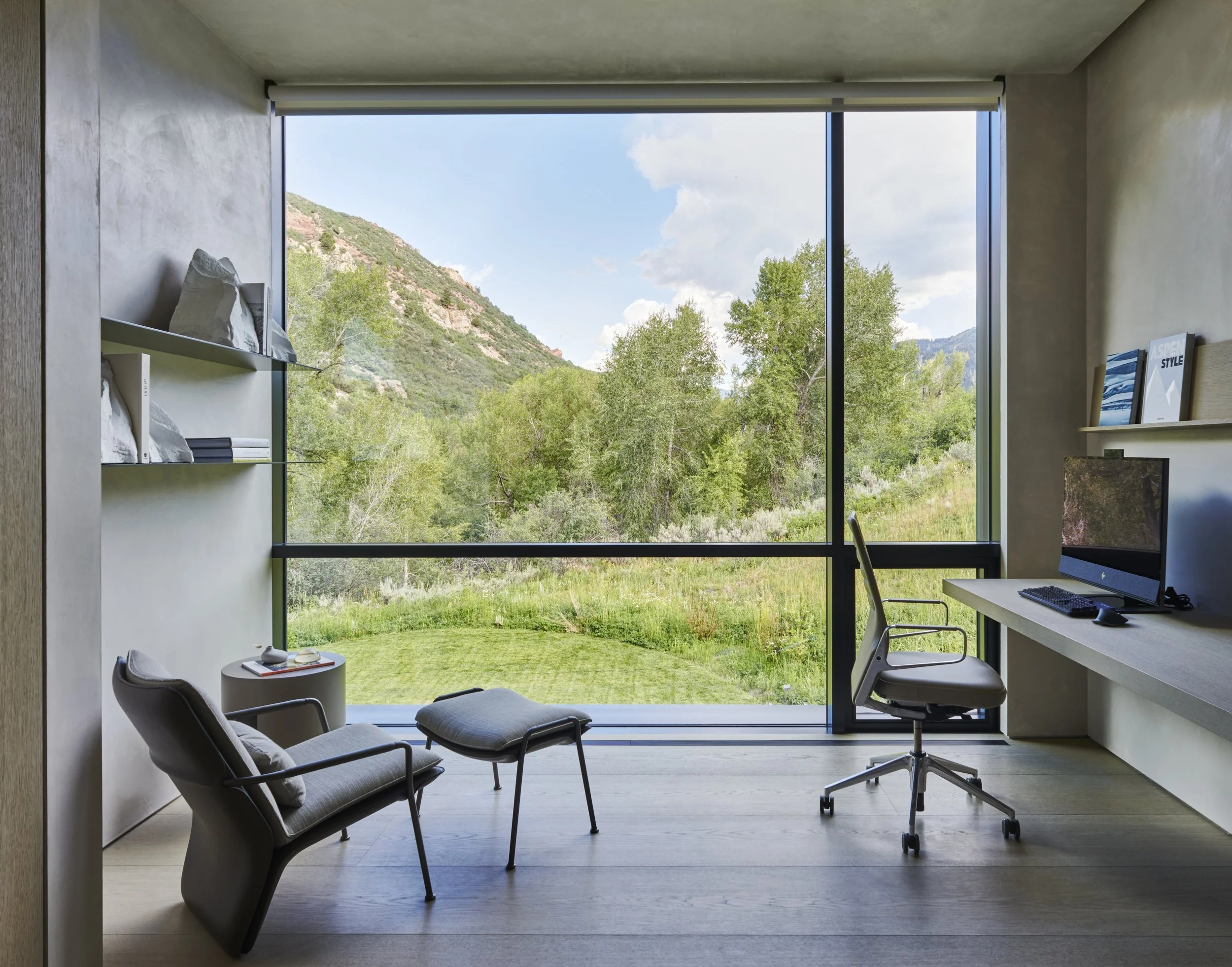 Modern interior room with large floor-to-ceiling window overlooking mountains and greenery, featuring a desk with a desktop computer, bookshelves, and lounge chairs.