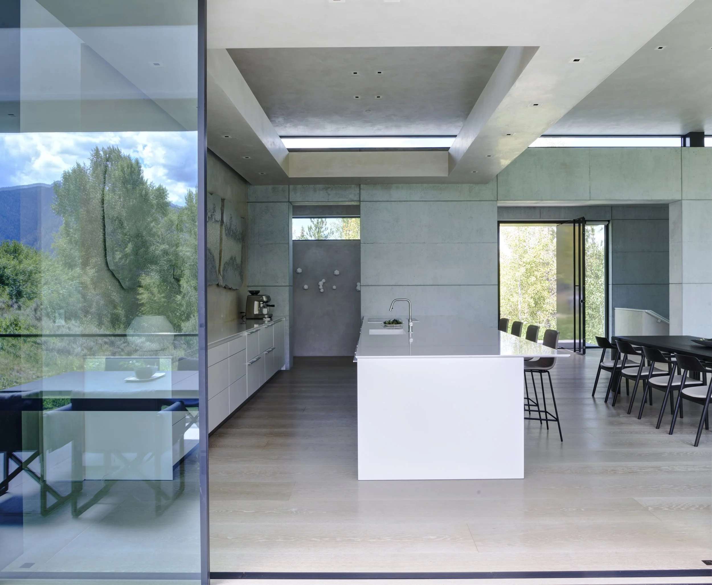 Interior view of a modern kitchen and dining area with concrete walls and ceiling, large windows showing a green landscape and blue sky, kitchen island with sink, and dining table with chairs.