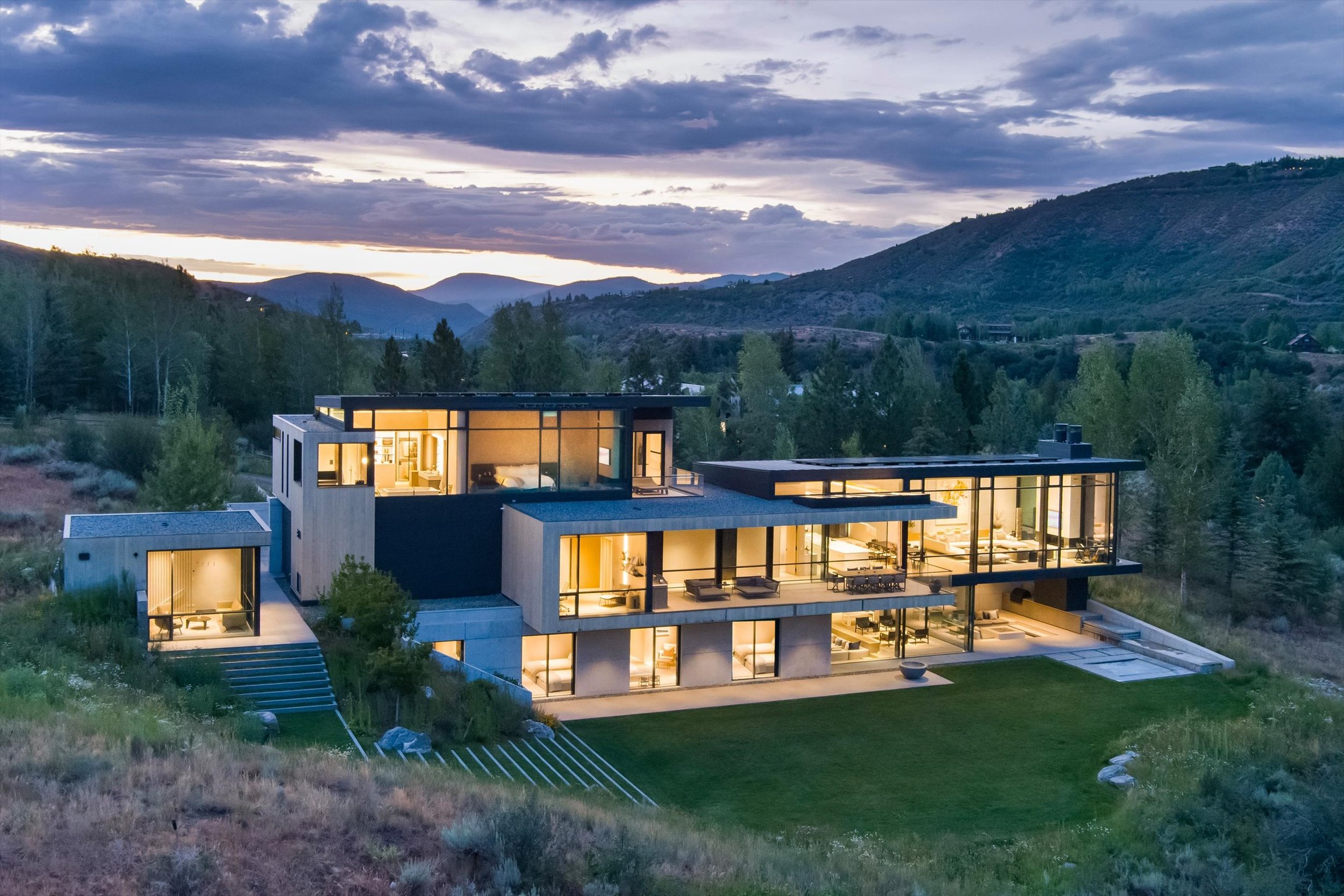 A modern multi-story house with large windows, illuminated from within, sitting on a hillside with a view of mountains and cloudy sky during dusk.