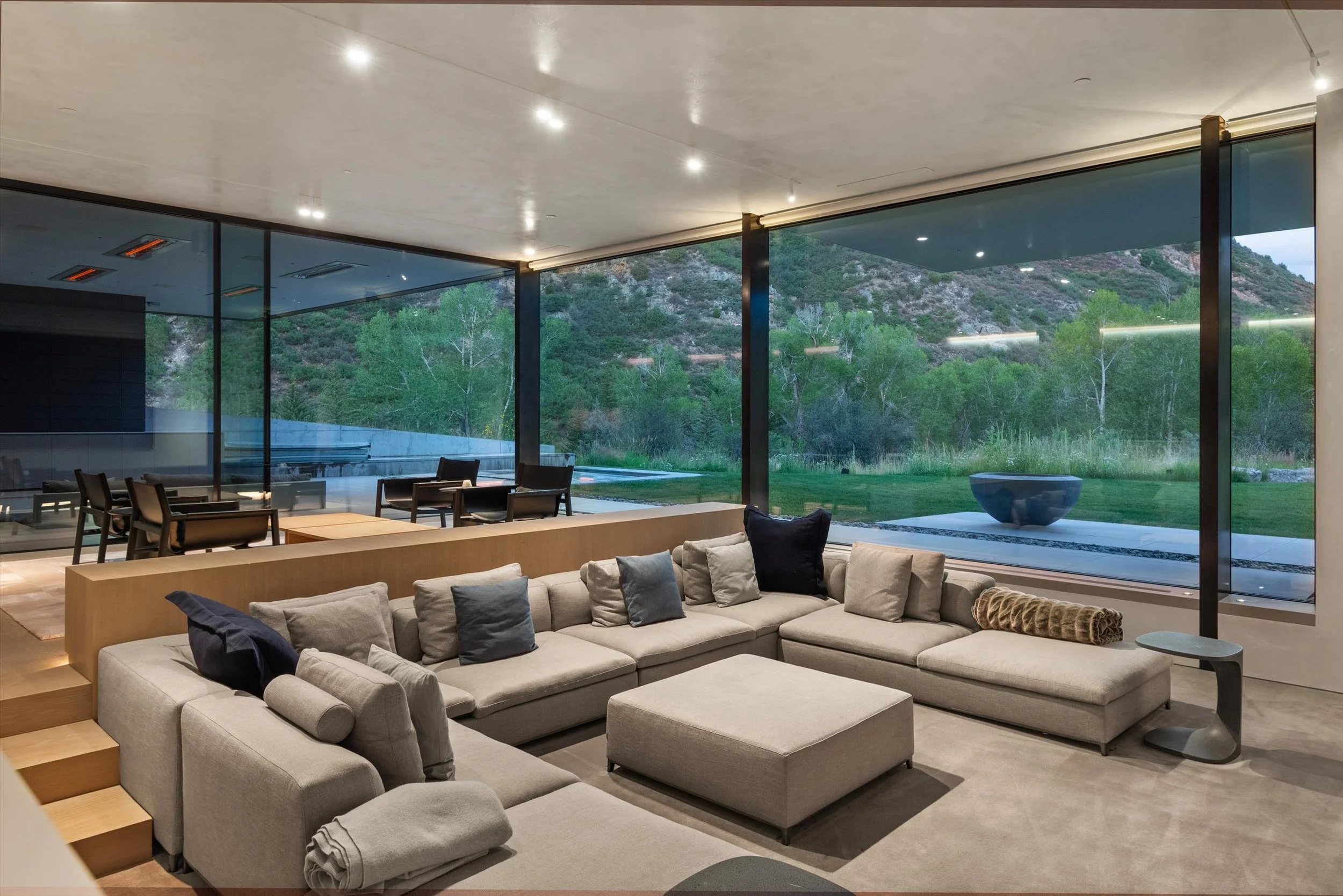 Modern living room with large glass windows overlooking a green mountainous landscape, featuring a beige sectional sofa with various pillows, meeting area with chairs, and a minimalist aesthetic.