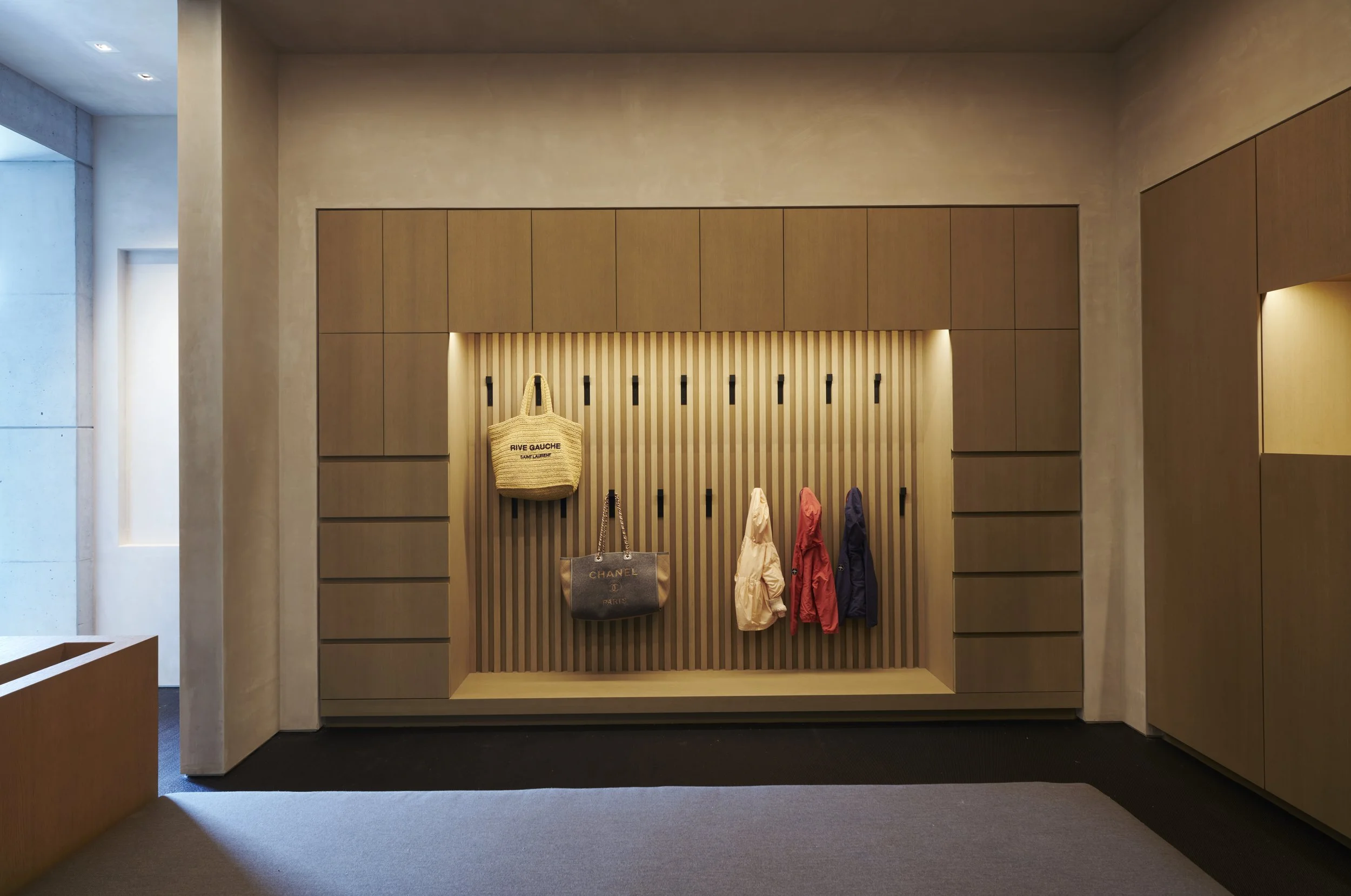 A built in modern nook featuring vertical slats. Several coats and handbags are hanging on hooks, including a beige tote bag with black lettering, a black Chanel tote, and various colorful jackets.
