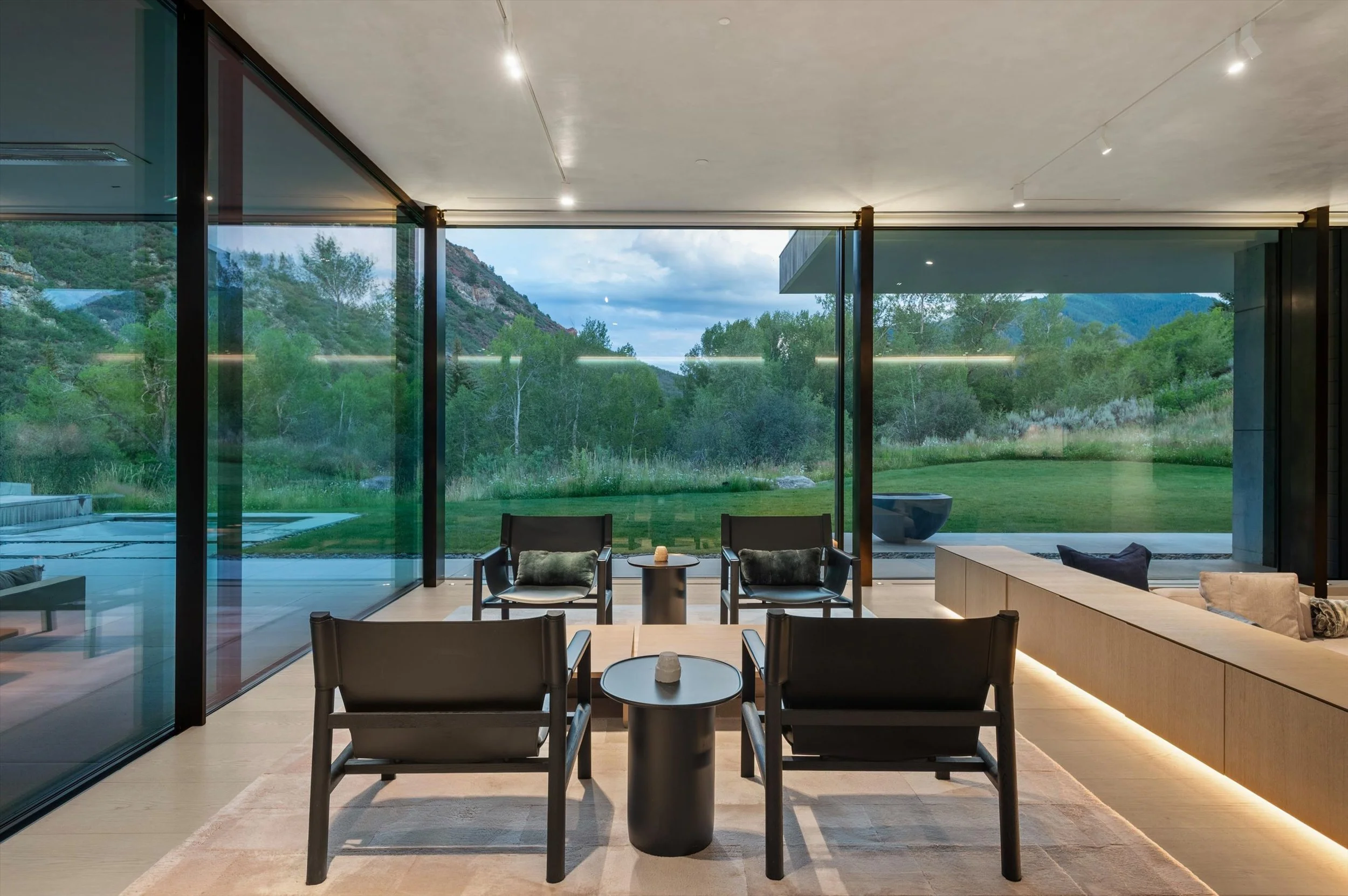 Modern living room with large glass windows showing a scenic mountain landscape, minimalist furniture, and ambient lighting.