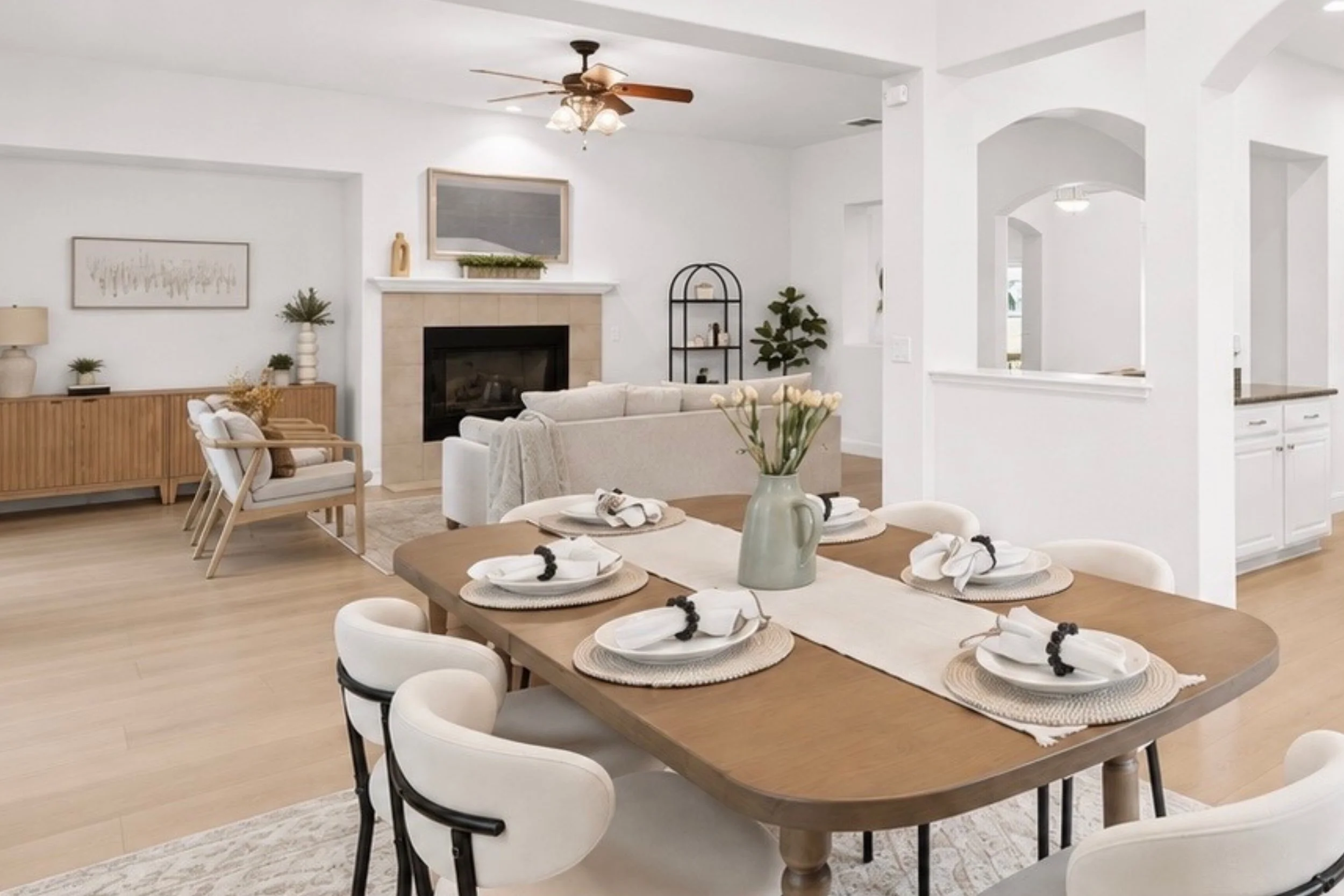 A dining room with a large wooden table set with white plates, napkins, and black napkin rings, surrounded by white and black chairs. In the background, a living area features a white sofa, a fireplace with a TV mounted above, and decorative plants. The room has white walls, a ceiling fan, and natural light.