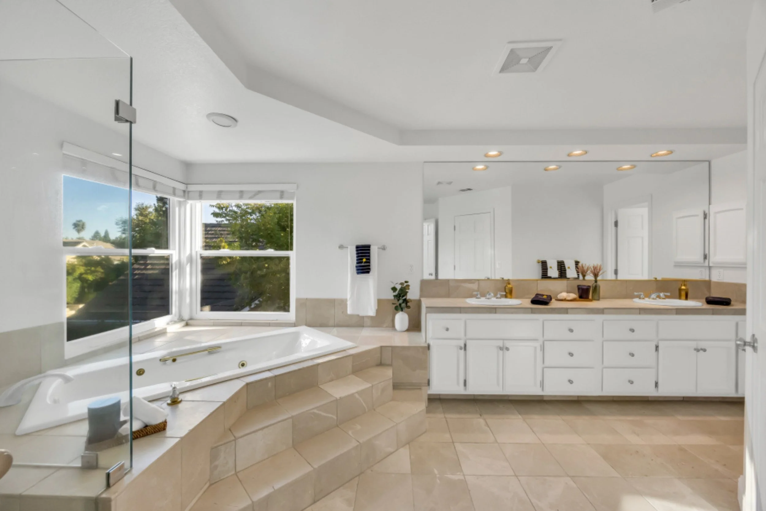 A spacious bathroom with a large window revealing trees and rooftops outside. The bathroom features a built-in bathtub with jet nozzles and a glass shower enclosure. There is a double sink vanity with white cabinets, a decorative vase, and a towel rack. The ceiling has multiple recessed lights and an air vent, and the walls and floor are tiled in beige and white.