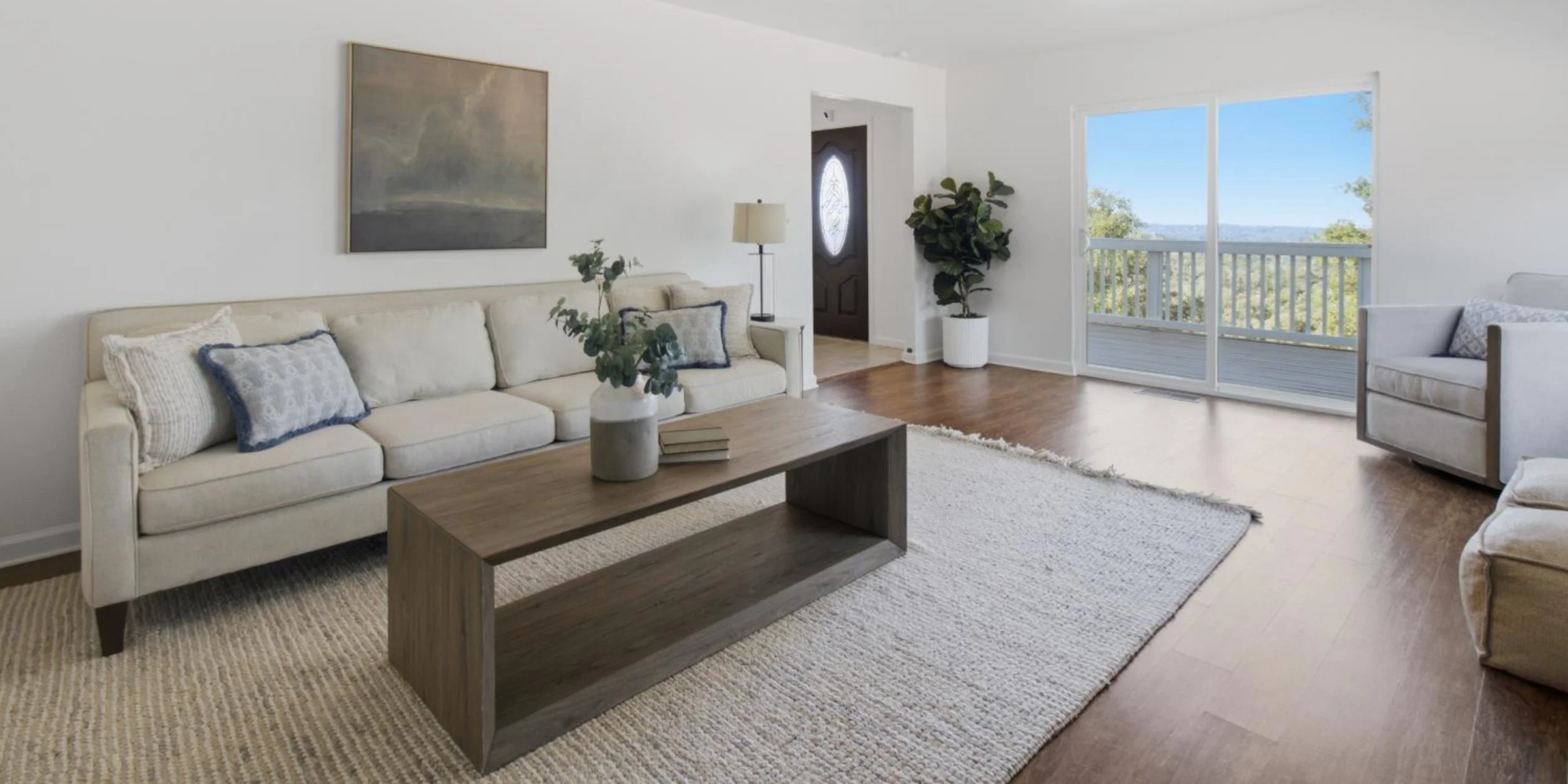 Living room with white sofa, armchair, wooden coffee table, large window with balcony, and artwork on white walls.