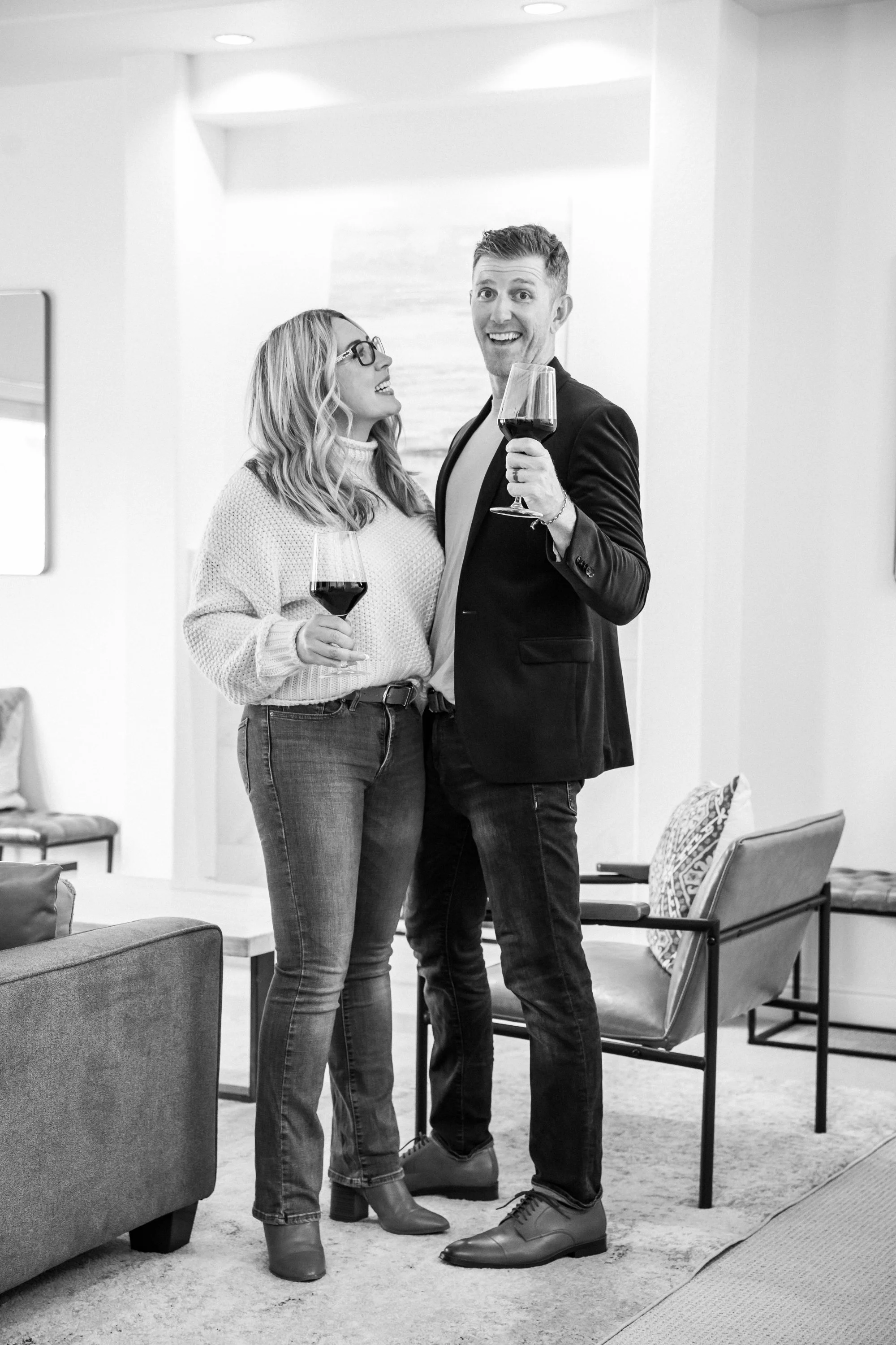 A man and a woman standing indoors, holding glasses of red wine, smiling and looking at each other. The woman has long wavy hair and glasses, wearing a sweater and jeans. The man has short hair, wearing a blazer and jeans. The room has modern furniture, including chairs and a rug.