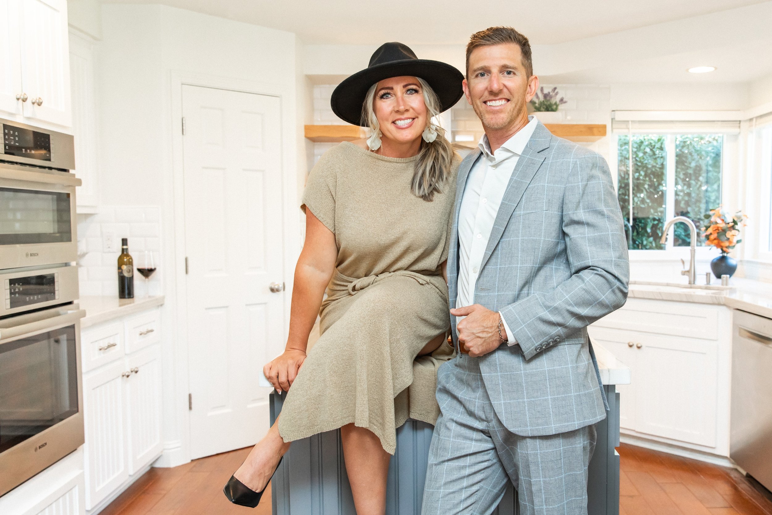 A smiling woman in a beige dress and black hat sitting on a kitchen kitchen counter with her arm around a smiling man in a gray checkered suit. The kitchen has white cabinets, a window, and a bottle of wine with a glass.
