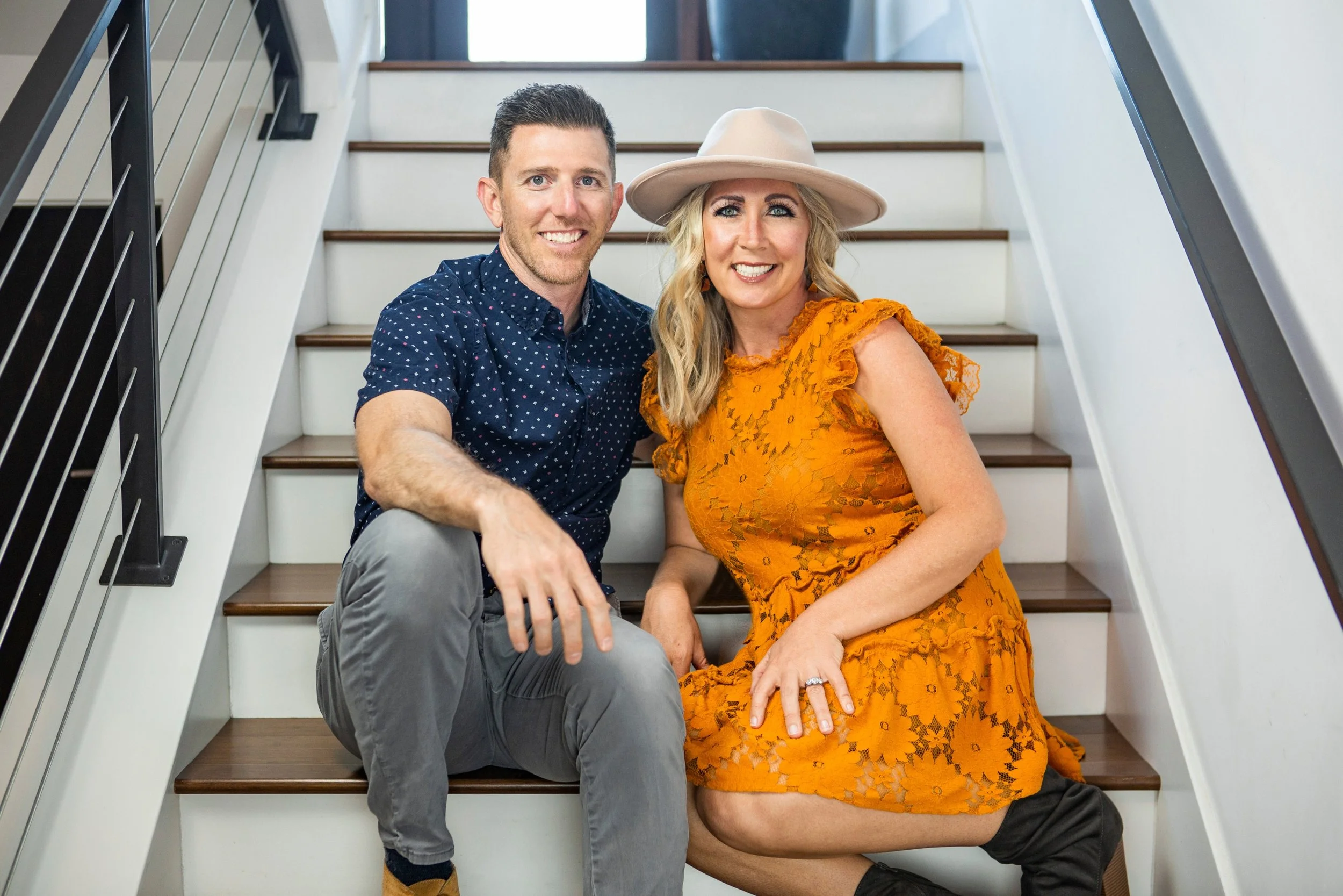 A man and woman sitting on stairs inside a house, smiling at the camera. The man has short dark hair, is wearing a blue polka-dot shirt, and grey pants. The woman has long blonde hair, is wearing a wide-brimmed beige hat and a bright orange lace dress, with one leg crossed over the other and wearing black boots.