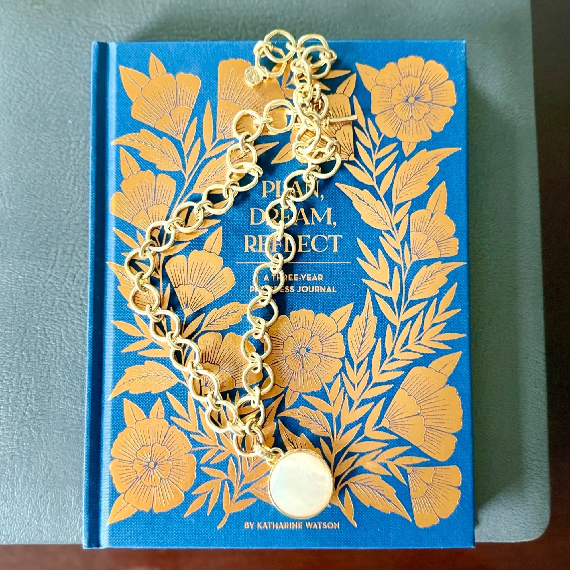 A blue hardcover book with gold floral patterns and gold text on the cover, partially obscured by a gold chain necklace with a large pendant, placed on a gray surface.