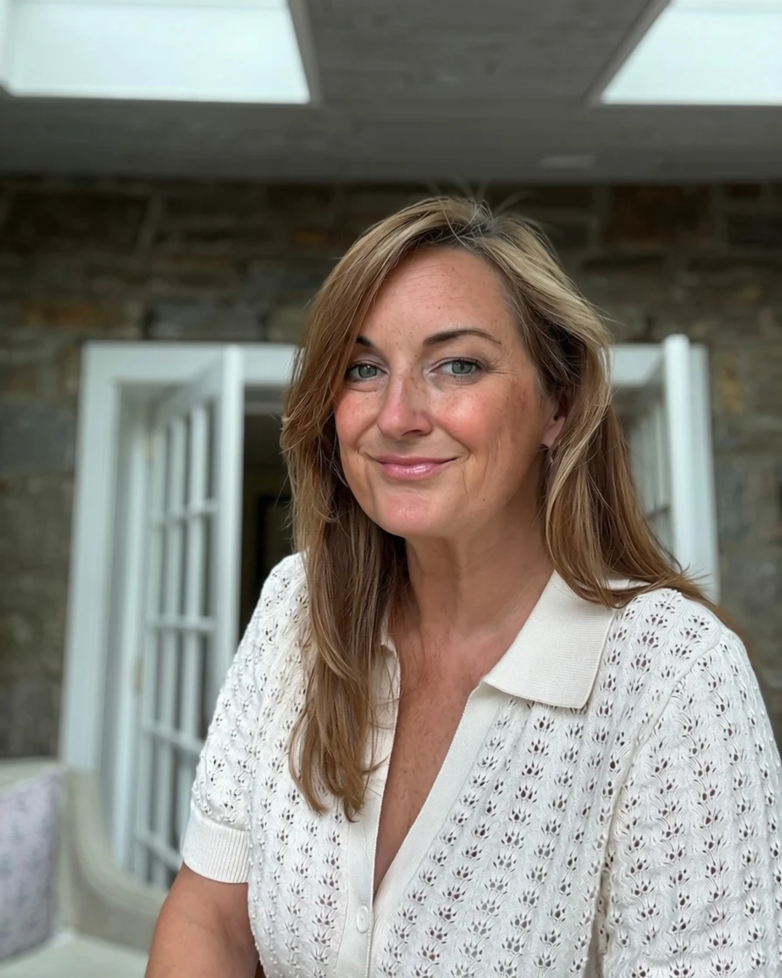 A woman with light brown, wavy hair and blue eyes smiling at the camera. She is wearing a white, patterned blouse with short sleeves. The background shows a stone wall and open white doors, with skylights above.