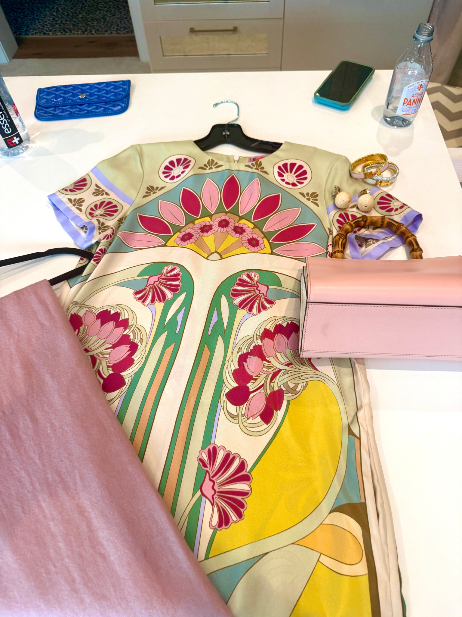 A colorful floral patterned dress on a hanger lying on a white table. Surrounding items include a pink fabric, jewelry, a green phone, water bottle, blue wallet, and a pink box.
