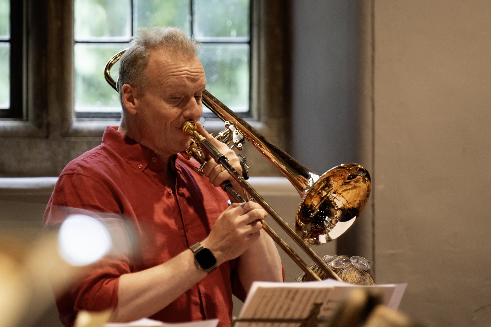 A man playing a trombone in a room with a window showing greenery outside.