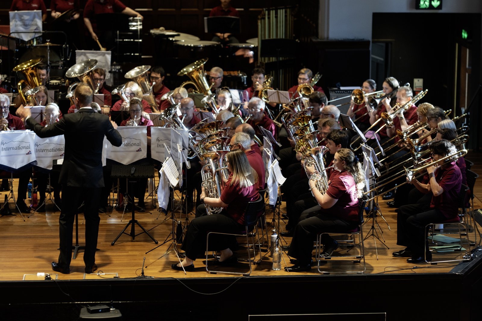 An orchestral concert with musicians playing various brass and percussion instruments, led by a conductor in a black suit, on a wooden stage with a dark background.