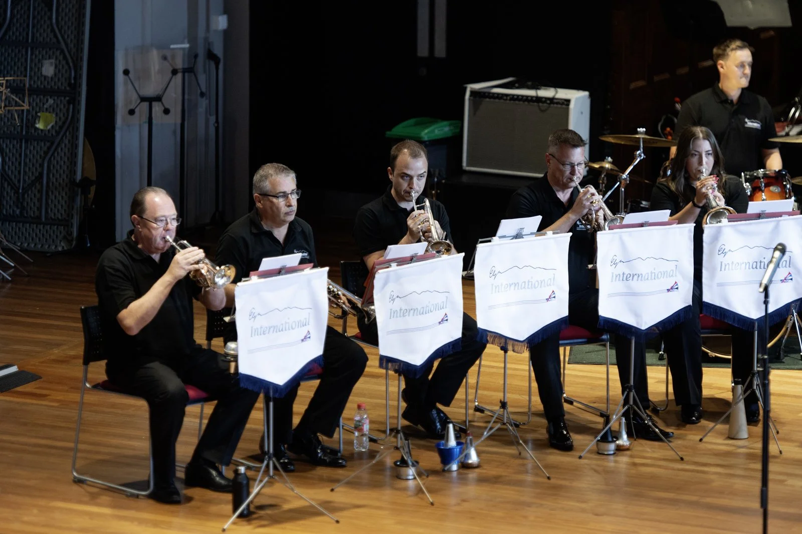 A group of five musicians sitting on a stage, playing trumpets, with sheet music in front of them. They are dressed in black, and there is a man in the background behind a drum set.