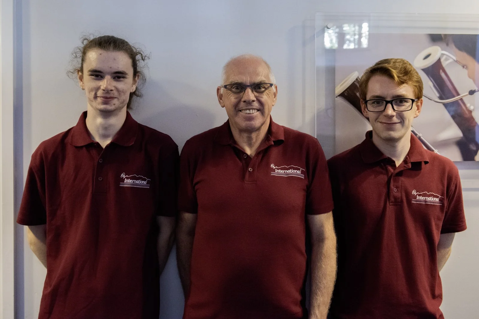 Three men in maroon polo shirts standing side by side, smiling, in front of a light-colored wall and glass display case.