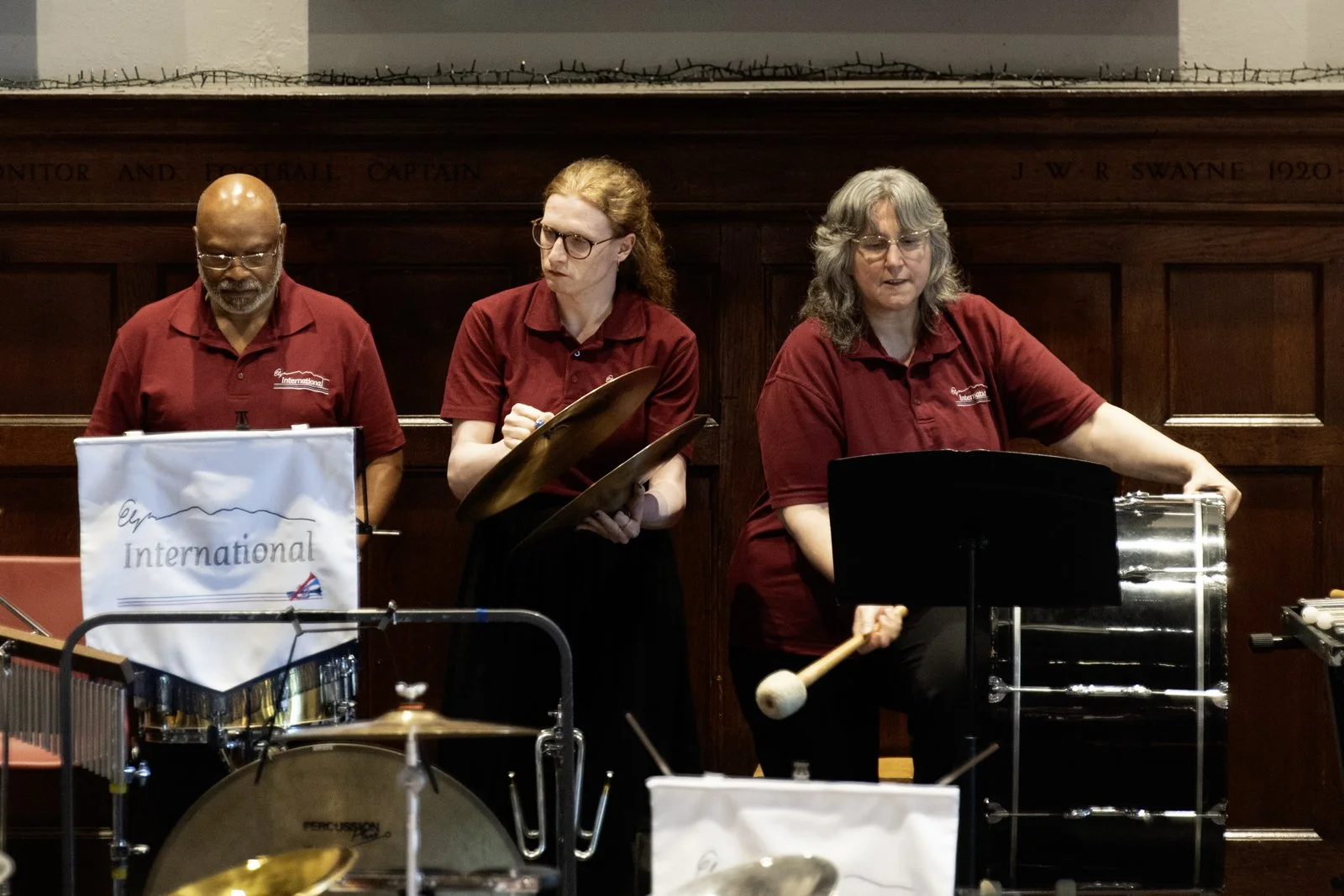 Three musicians in red shirts performing with percussion instruments in a wood-paneled room.