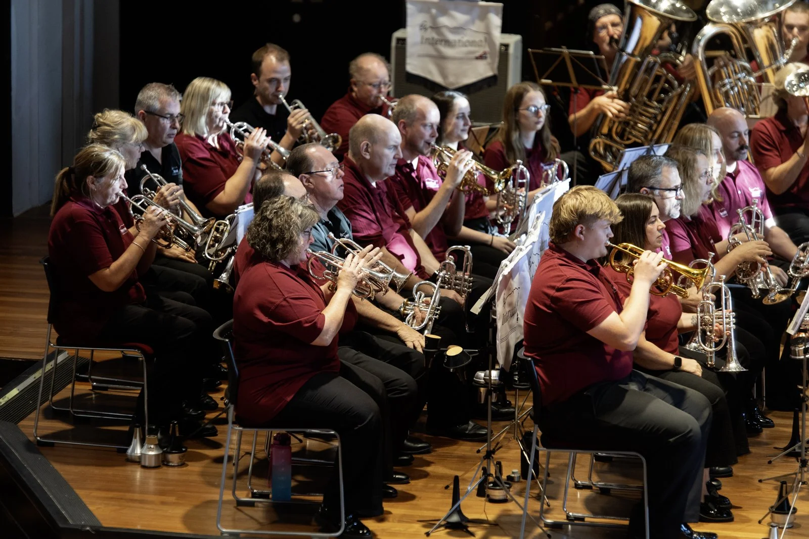 A large band of musicians in maroon shirts playing wind instruments on stage during a concert.
