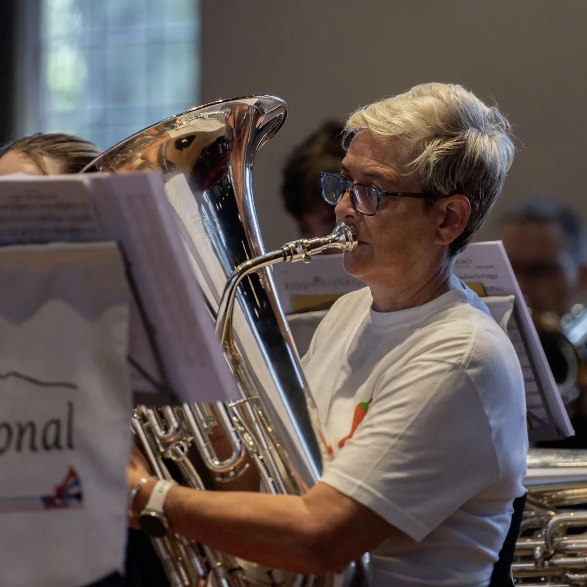 An older woman with short blonde hair and glasses playing a tuba during a concert or rehearsal.
