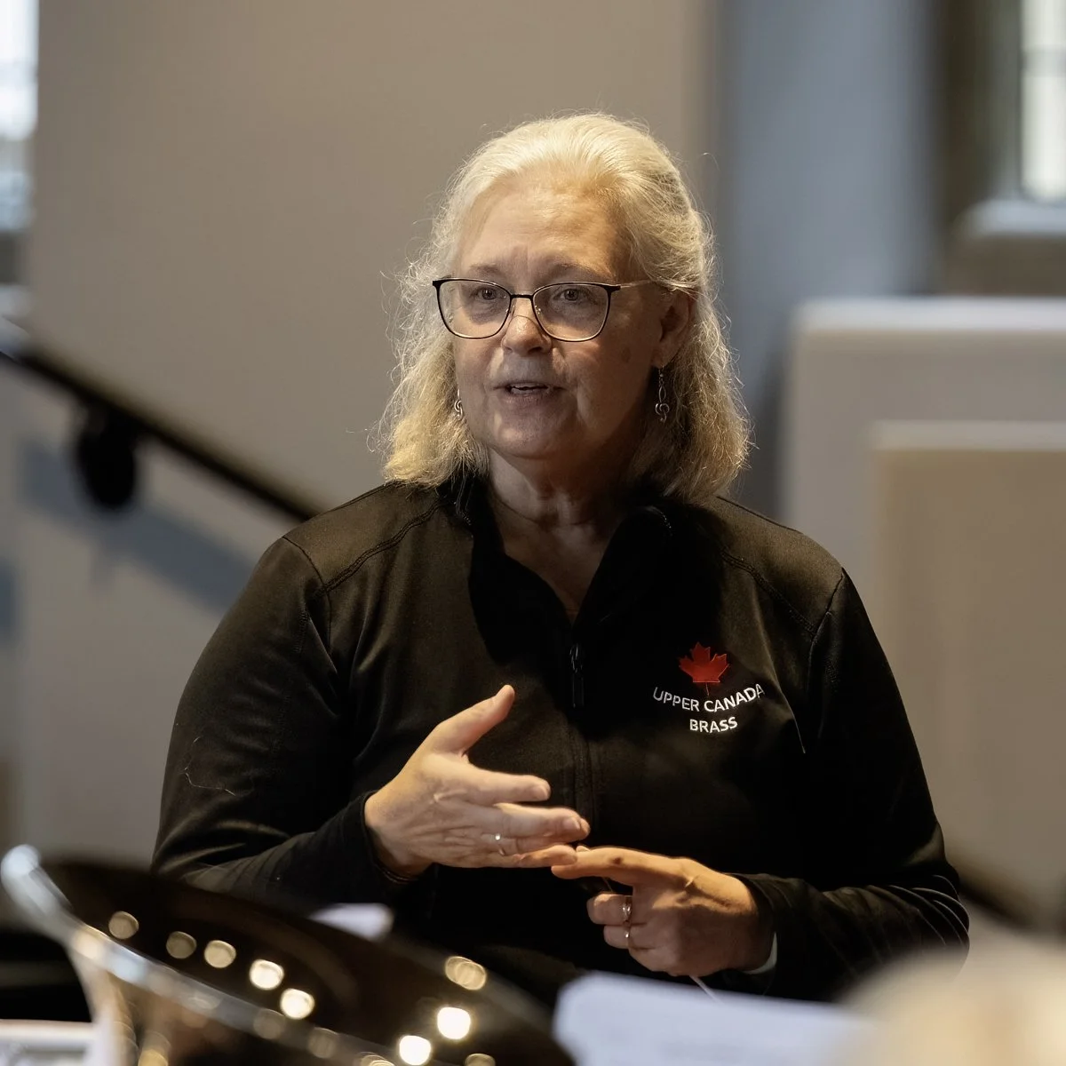 A woman with gray hair and glasses, wearing a black jacket with a red maple leaf and text 'Upper Canada Brass,' speaking and gesturing with her hands in a room with a window and a white object in the background.