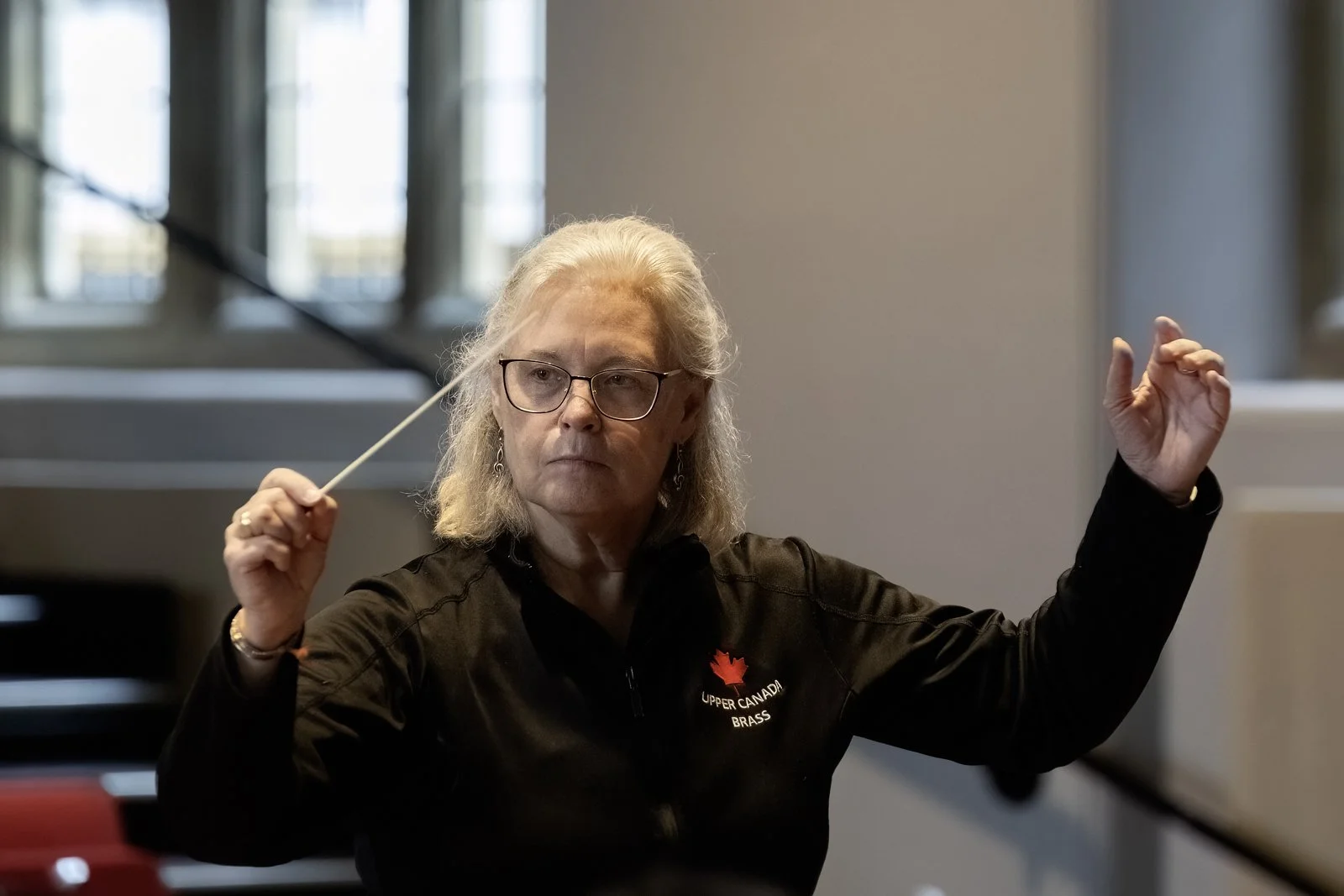 A woman with long gray hair and glasses conducting an orchestra, wearing a black jacket with an 'Upper Canada Brass' logo, using a conductor's baton in a well-lit indoor setting.