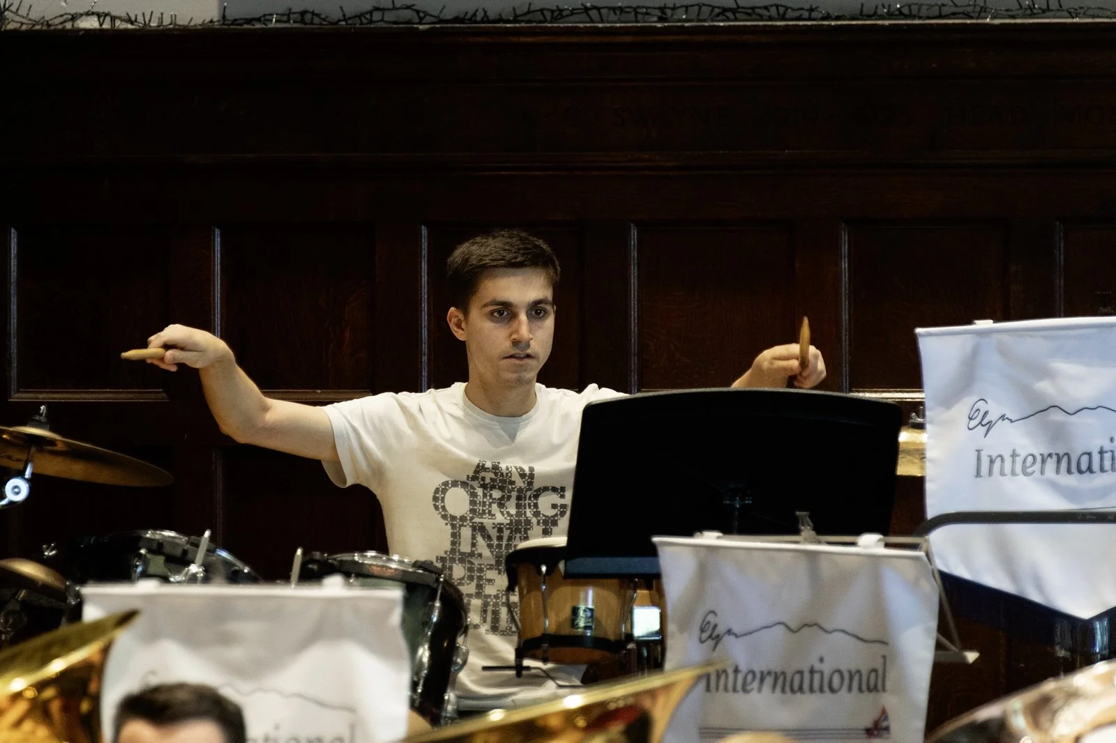 Young man playing drums during an indoor concert with a wood-paneled wall background and music stands with 'Eyp' and 'International' written on them.