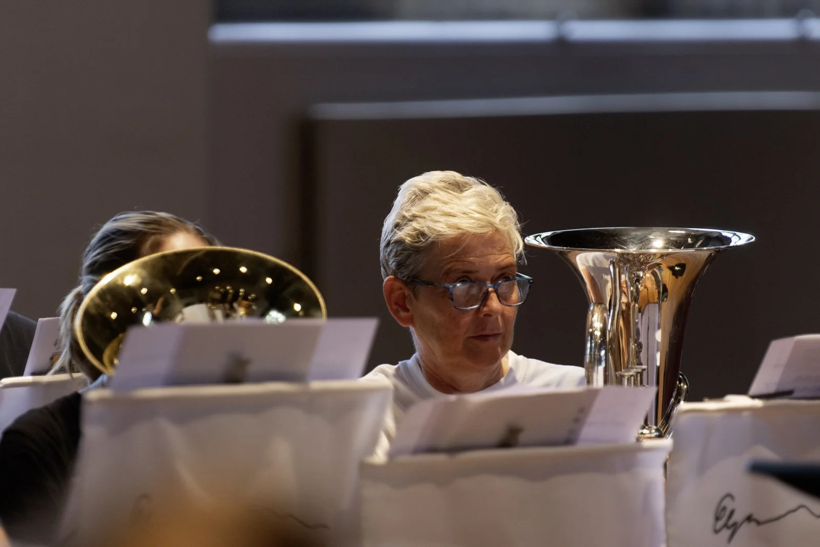 A person with short gray hair and glasses sitting among music stands with sheet music, in a room with a plain background, during a musical performance or rehearsal.