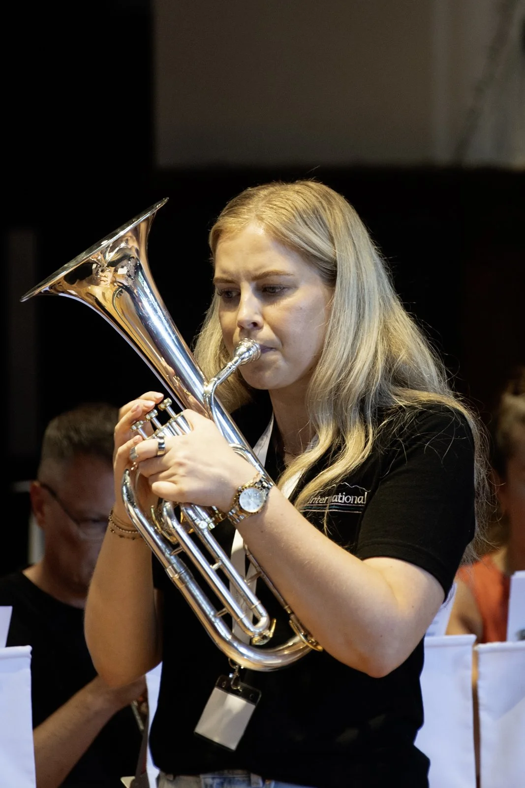 A woman with blonde hair playing a silver euphonium, focused on her music, in a band or orchestra setting.