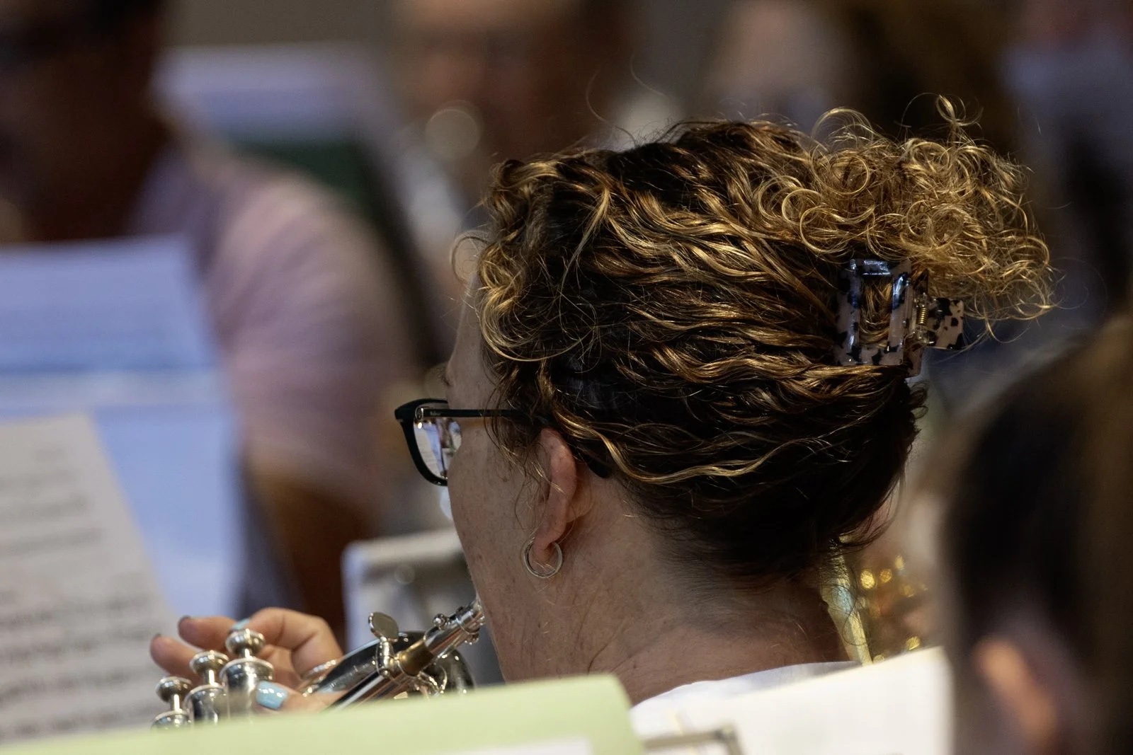 A woman with curly hair in a hair clip, wearing glasses and earrings, holding a trumpet, with blurred sheet music and other people in the background.