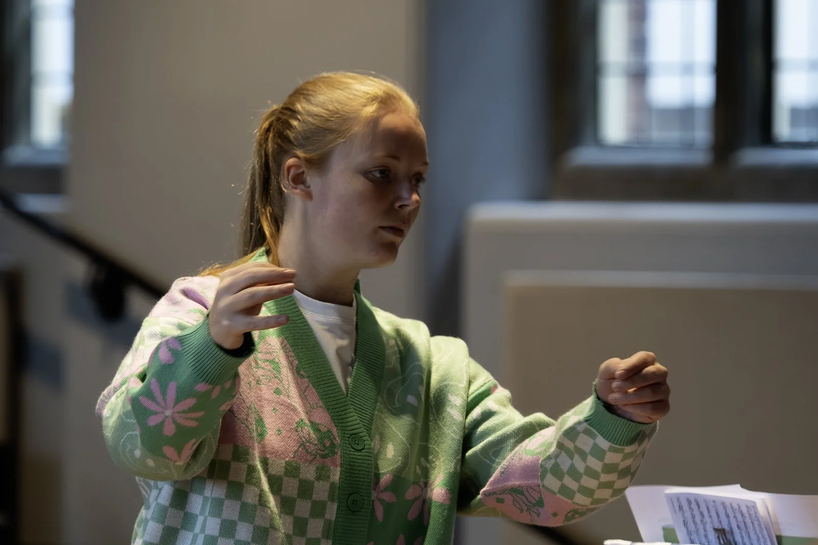 A young girl with red hair, wearing a patterned green and pink sweater, standing indoors near a staircase, gesturing as she looks at a document or paper.
