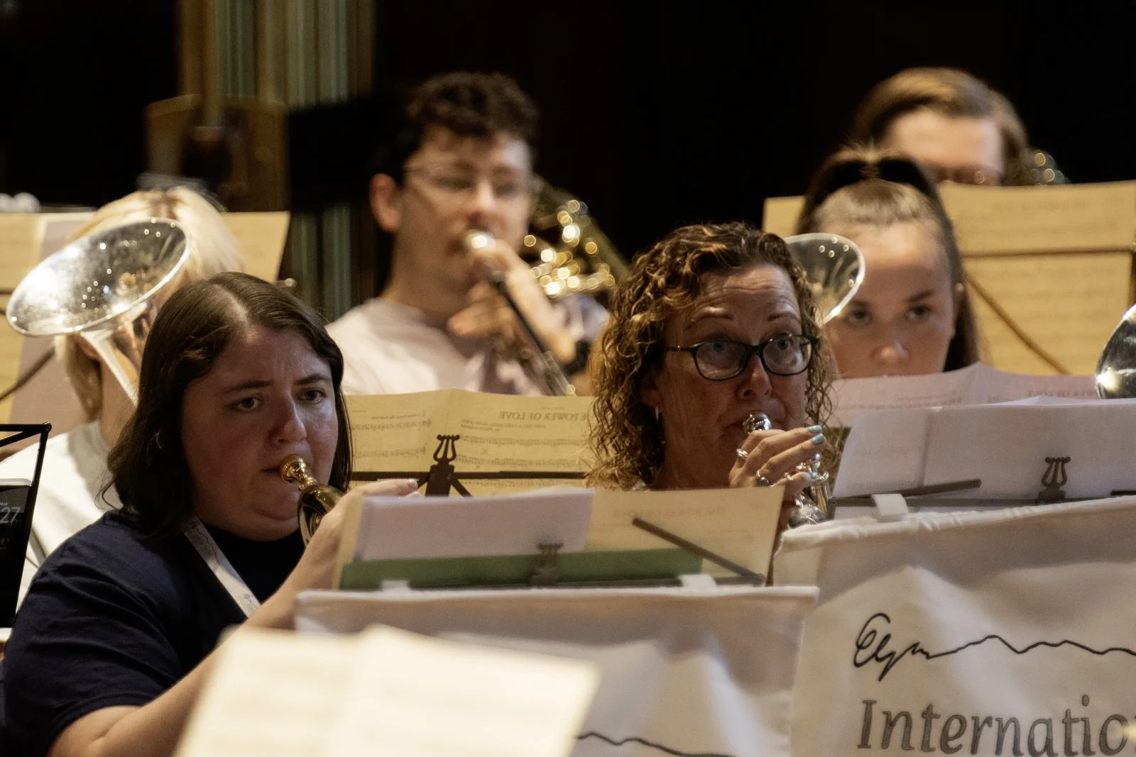 A group of people playing wind instruments, including flutes, in an orchestra setting with music stands and sheets in front of them, on a stage.