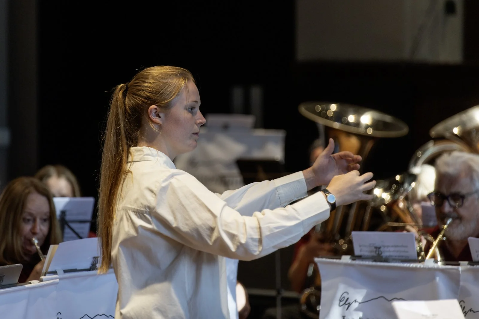 A young woman conducting a band, with musicians playing brass instruments in a concert setting.