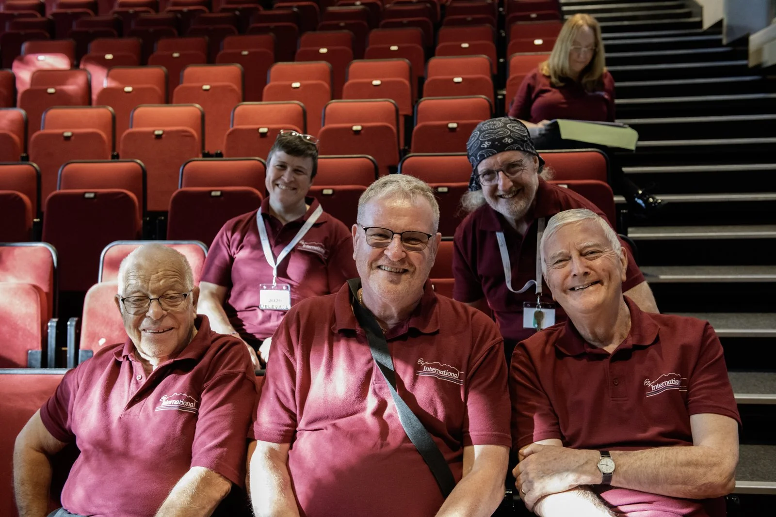 Group of six smiling people wearing maroon shirts sitting and standing in an auditorium with red seats.