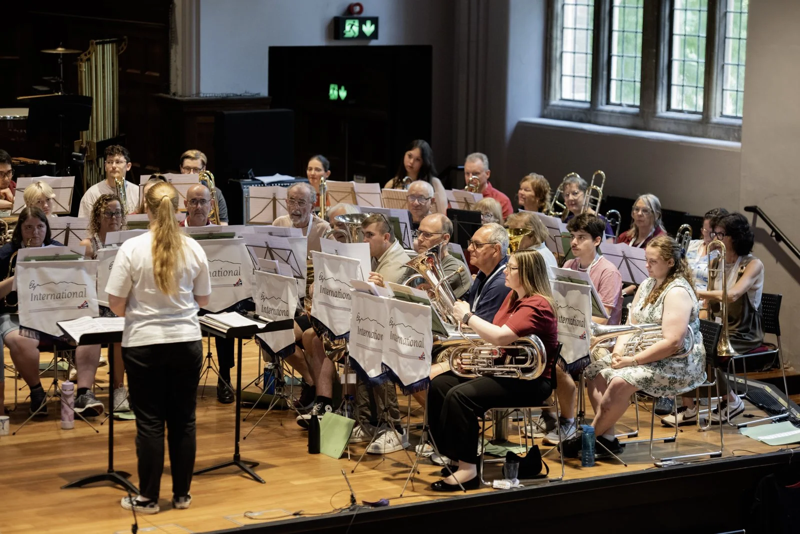 An indoor concert hall with a large ensemble of musicians seated and preparing for a performance, with a conductor standing in front of them.