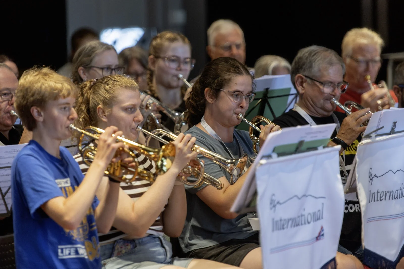 Group of people playing trumpets during a band rehearsal or performance, seated with music stands in front of them.