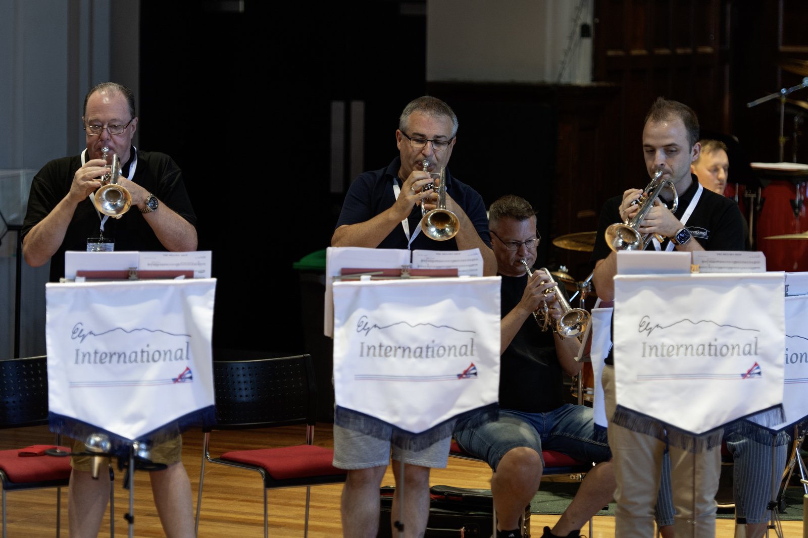 Group of four musicians playing trumpets during a band performance, seated behind music stands labeled 'International,' in a room with a wooden floor and dark walls.
