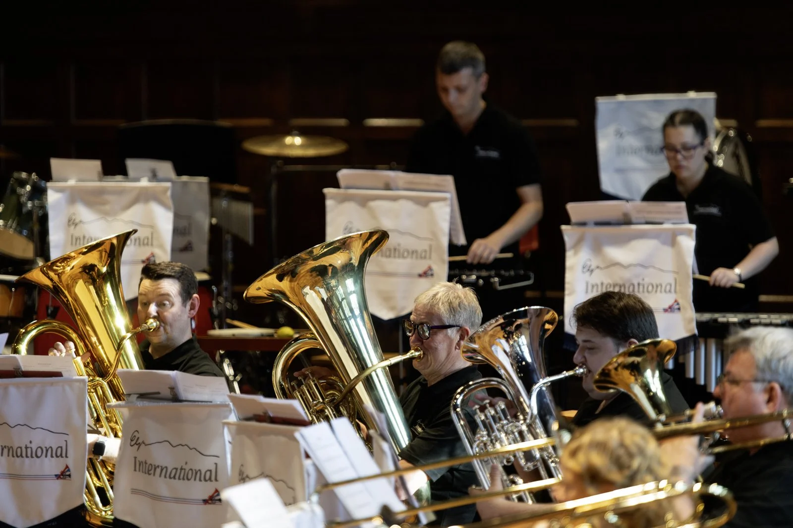 Orchestra performing on stage with musicians playing brass instruments and percussion, reading music from stands with banners labeled 'International'.